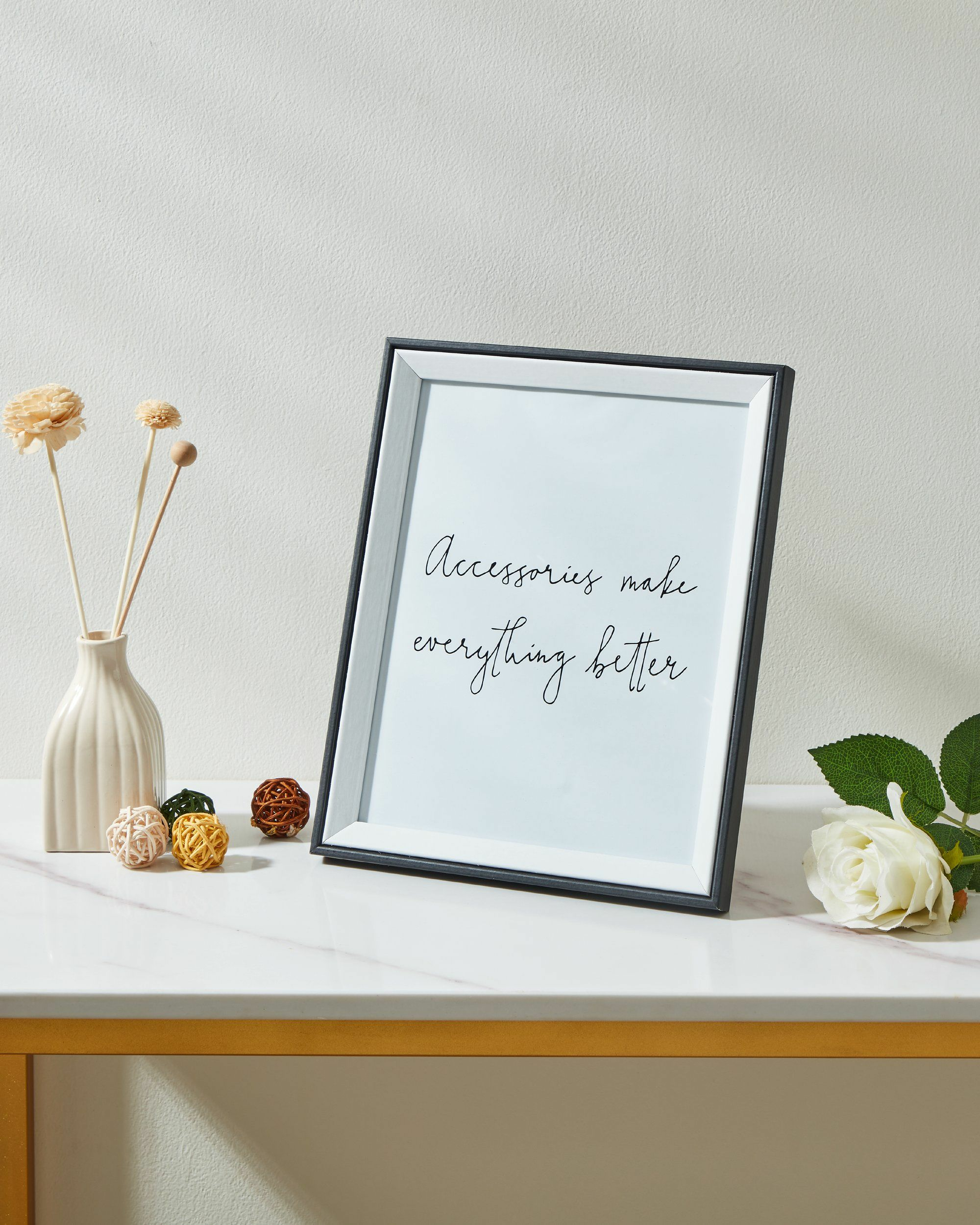 Une image encadrée avec du texte, un vase avec des fleurs séchées et une rose blanche sur une table en marbre.