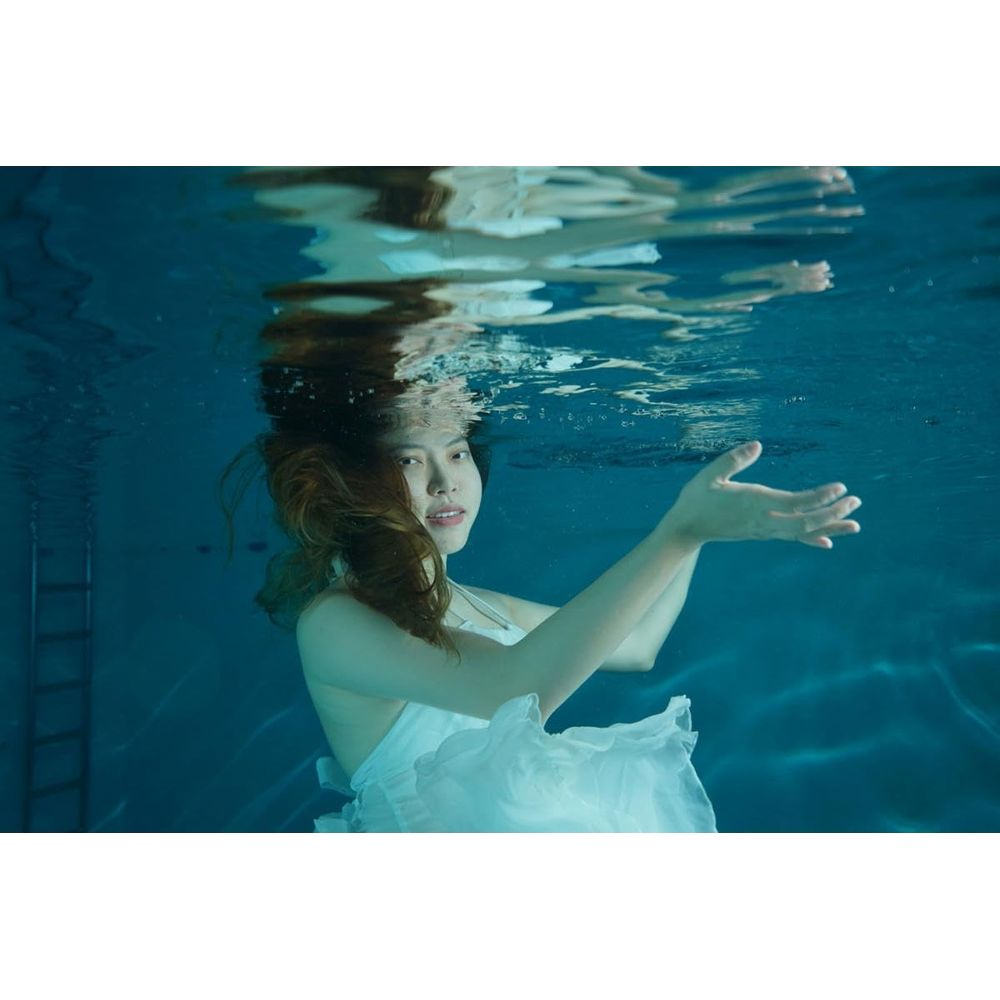 Une femme en robe blanche sous l'eau dans une piscine. Ses cheveux flottent autour d'elle.