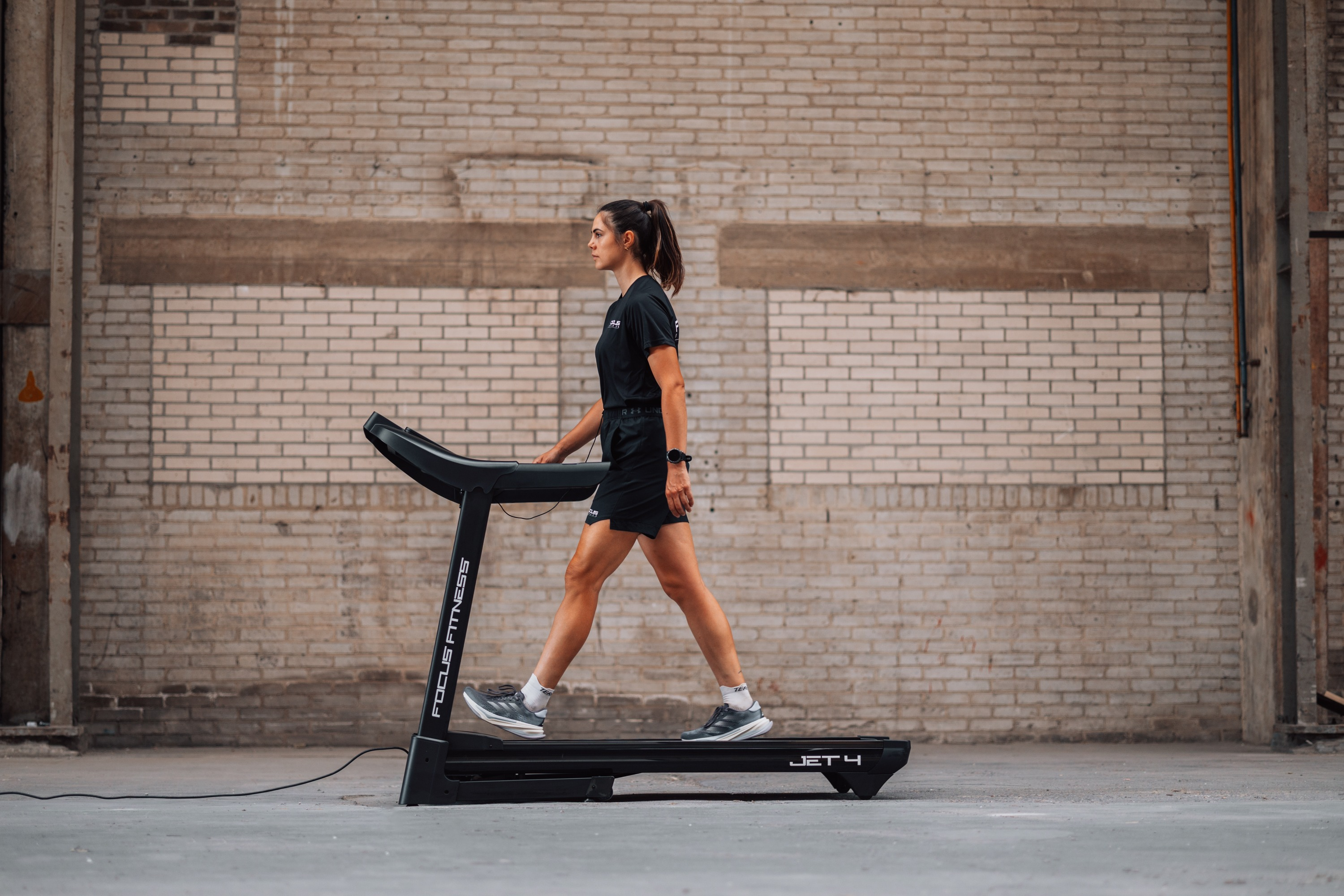 A woman in black walks on a treadmill in front of a brick wall.