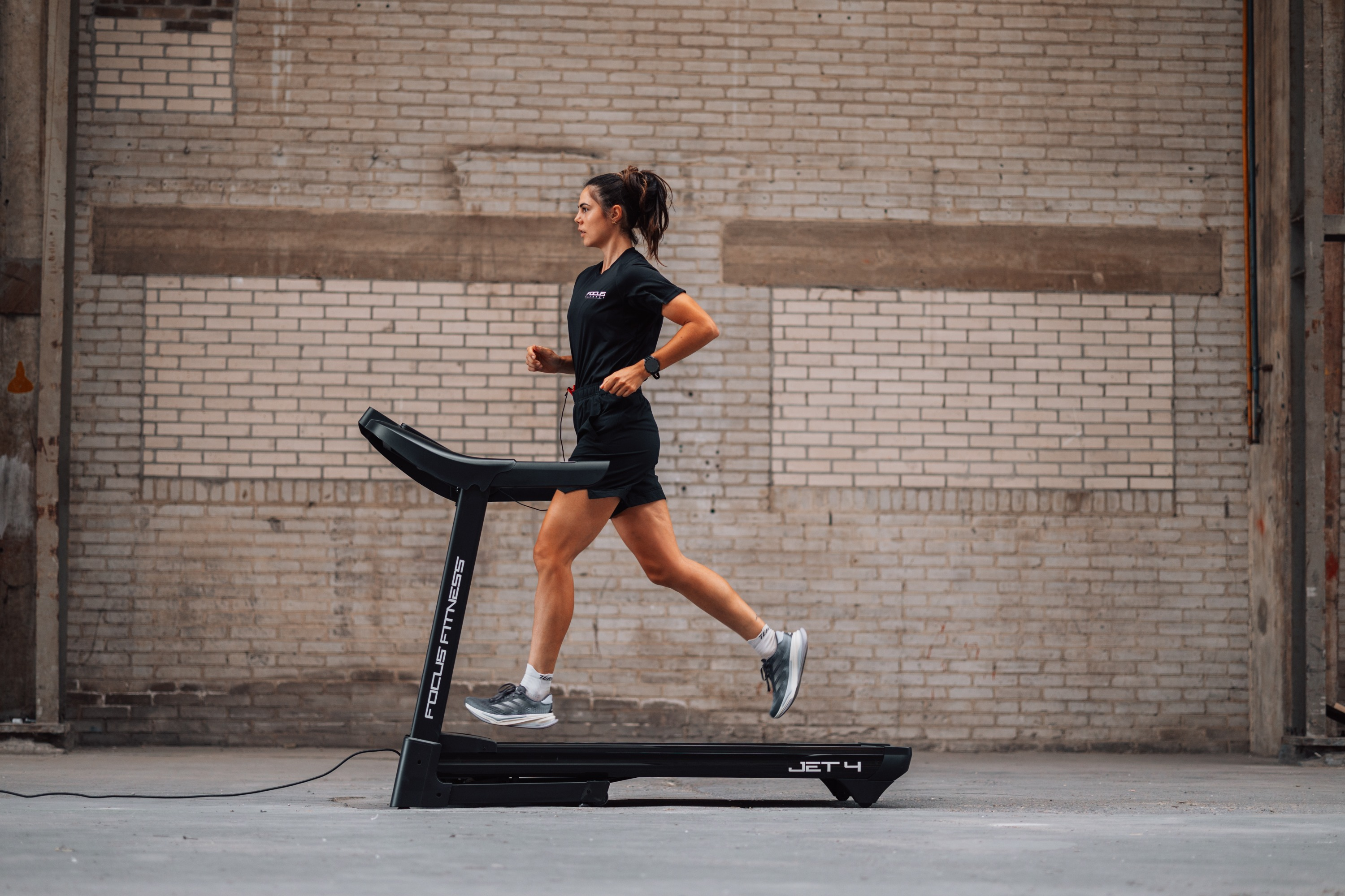 Woman in black runs on treadmill in front of brick wall