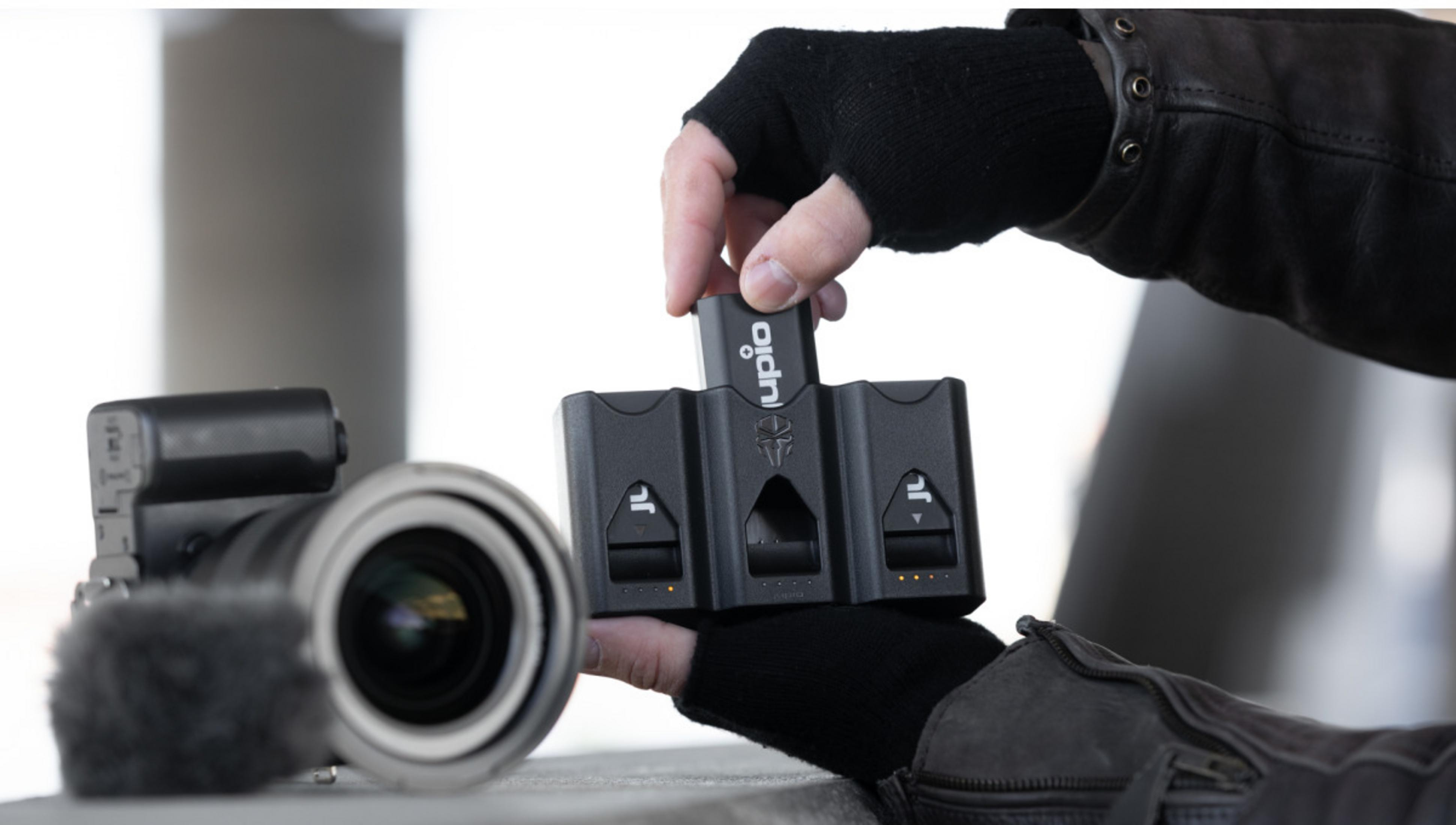 Una mano con guanto nero inserisce una batteria in un caricabatterie accanto a una fotocamera.