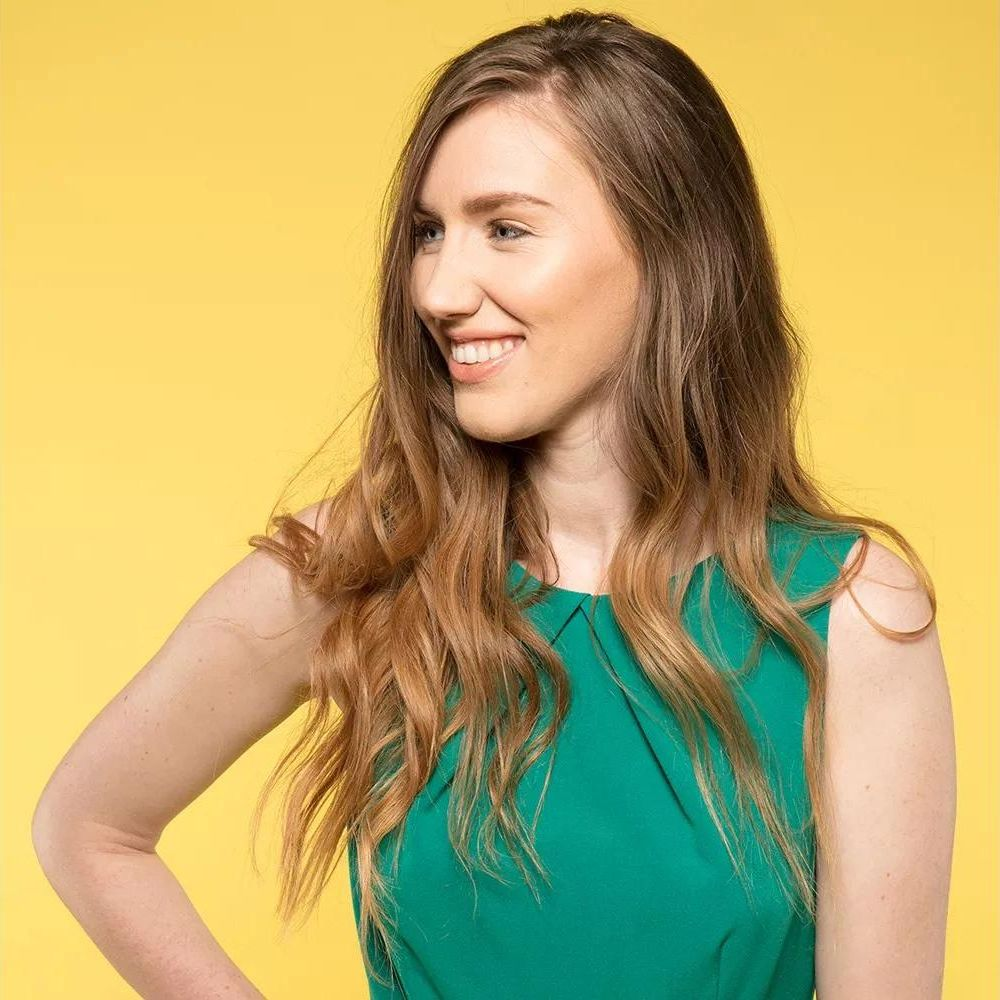 Une femme aux longs cheveux bruns sourit sur fond jaune.