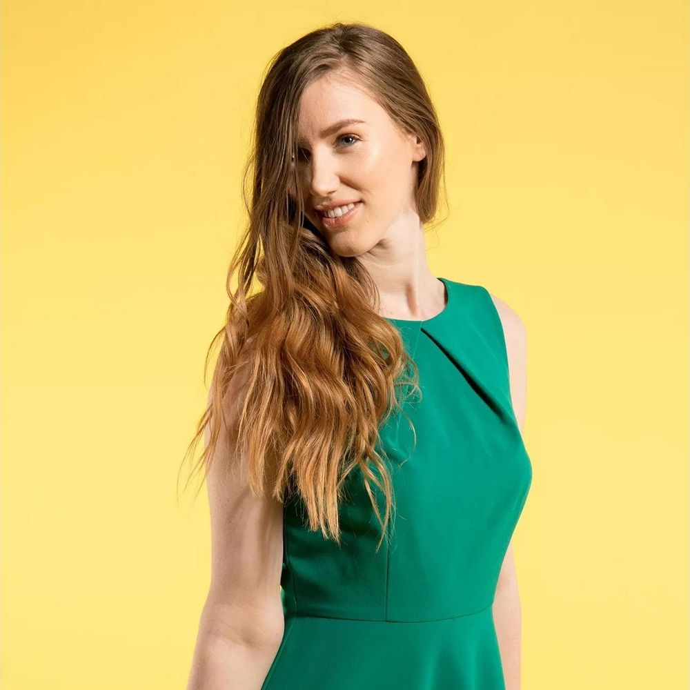 Une femme aux longs cheveux bruns porte une robe verte sur fond jaune.