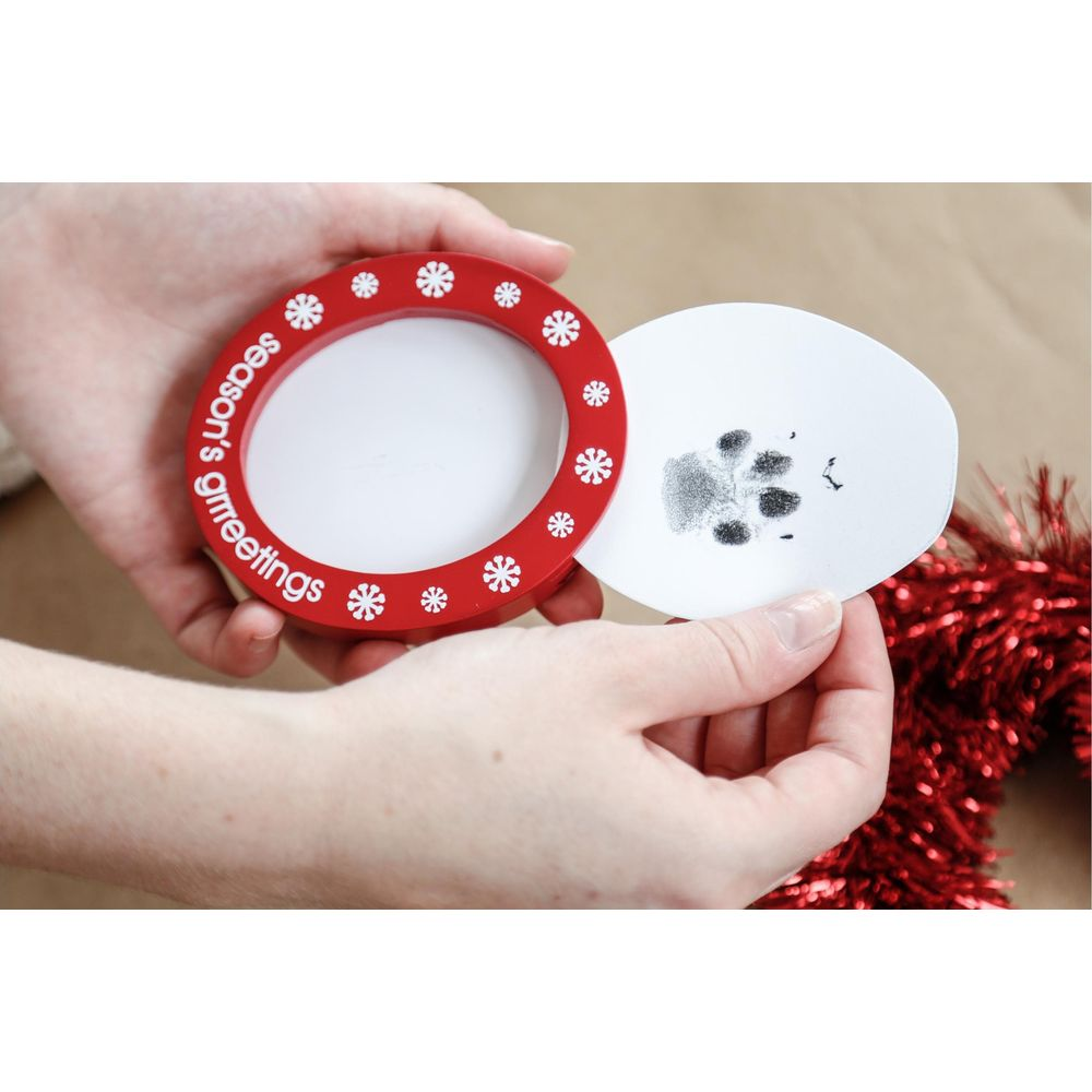 Des mains tiennent un cadre rouge 'Season's Greetings' et une carte avec une empreinte de patte.