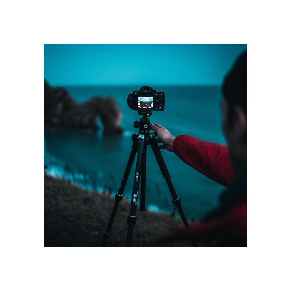 Une personne en veste rouge installe un appareil photo sur trépied, face à l'océan.