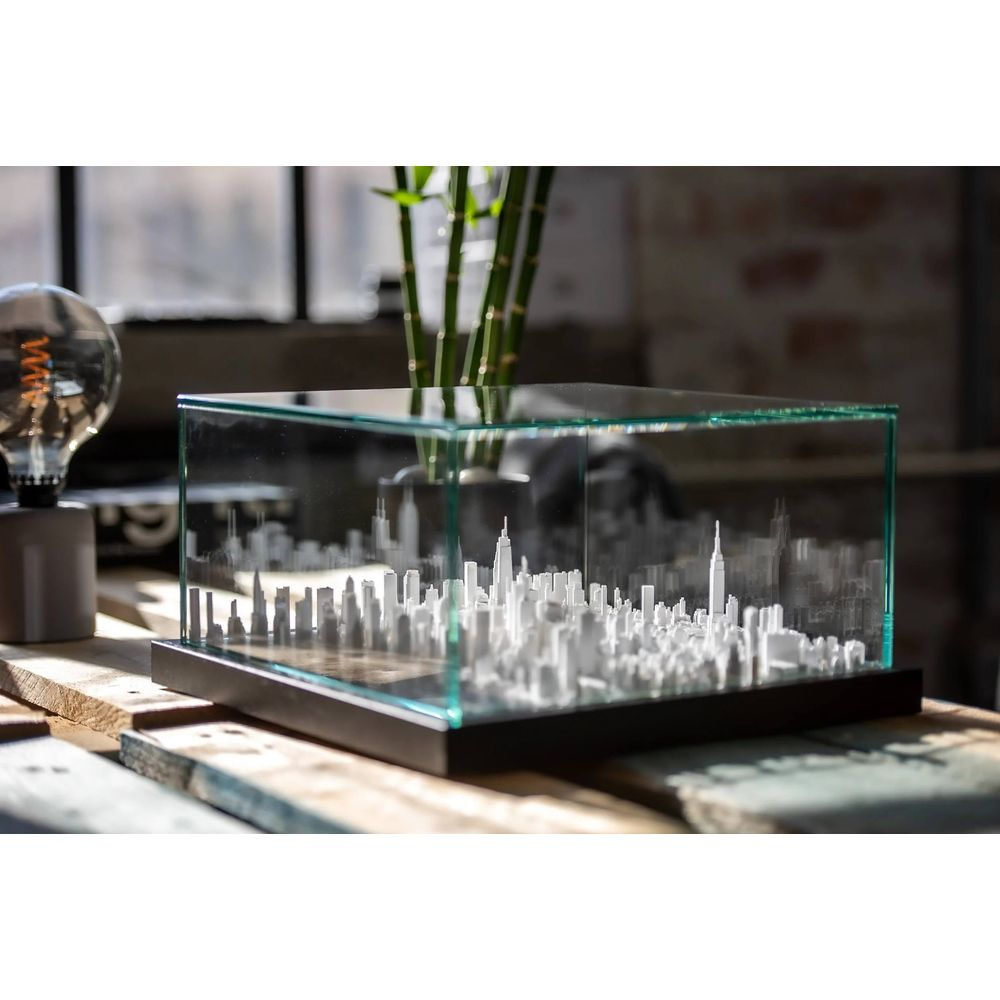 Une maquette de ville 3D blanche dans une vitrine en verre sur une table en bois, avec une ampoule.