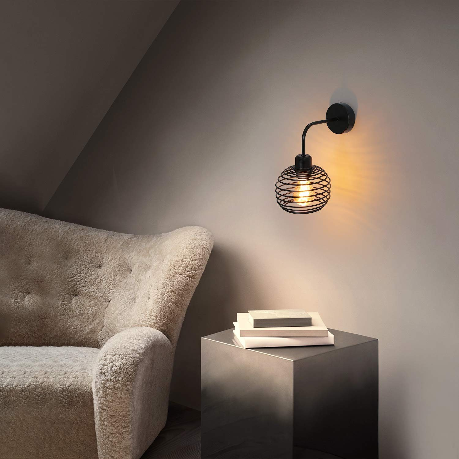 Un sillón acogedor junto a un cubo metálico con libros. Una lámpara de pared negra brilla cálidamente.