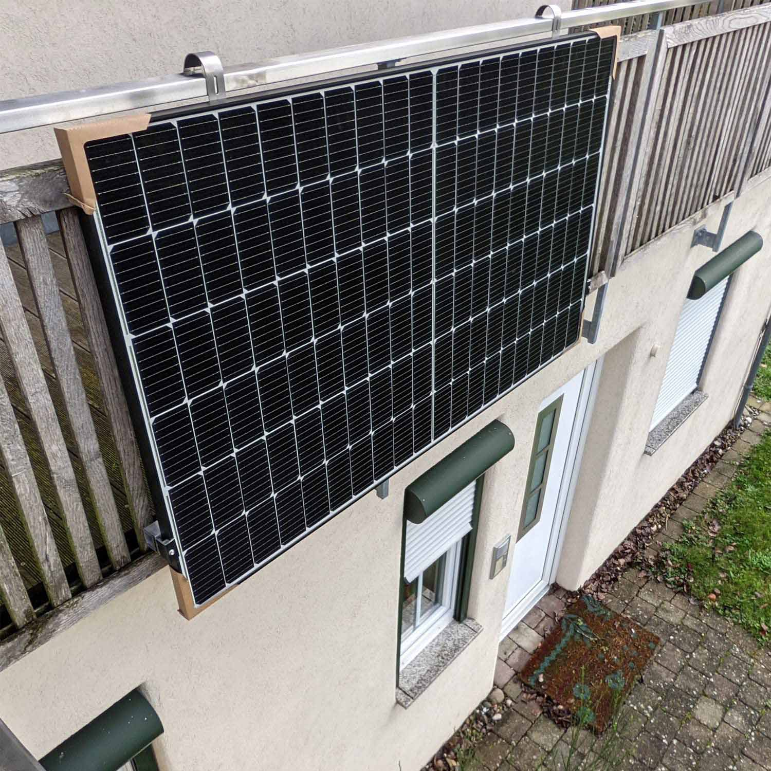 Ein schwarzes Solarpanel ist an einem Balkongeländer befestigt. Daneben befindet sich eine Hauswand mit einem Fenster und einer Tür.