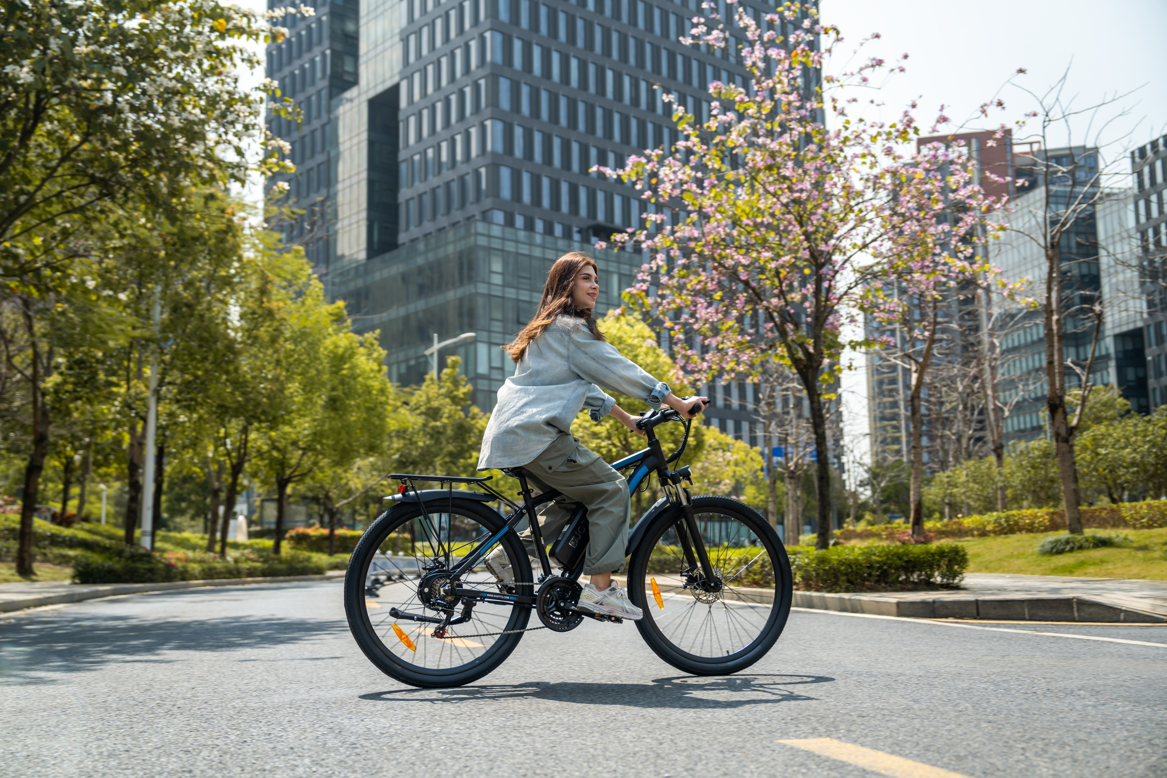 Eine Frau fährt ein E-Bike auf einer Straße mit Bäumen und modernen Gebäuden im Hintergrund.