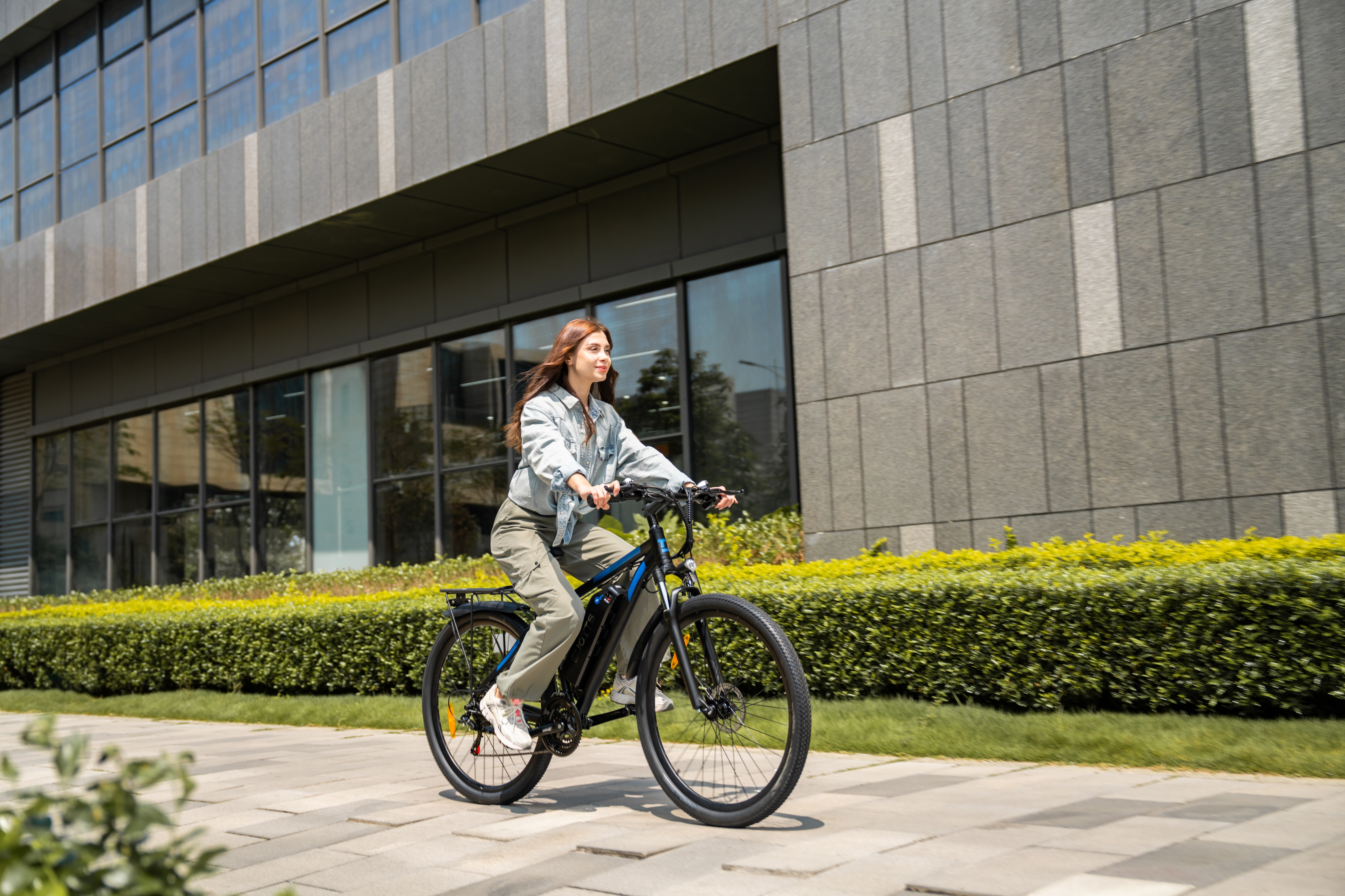 Eine Frau fährt ein schwarzes E-Bike vor einem modernen Gebäude mit Glasfenstern und grünen Büschen.