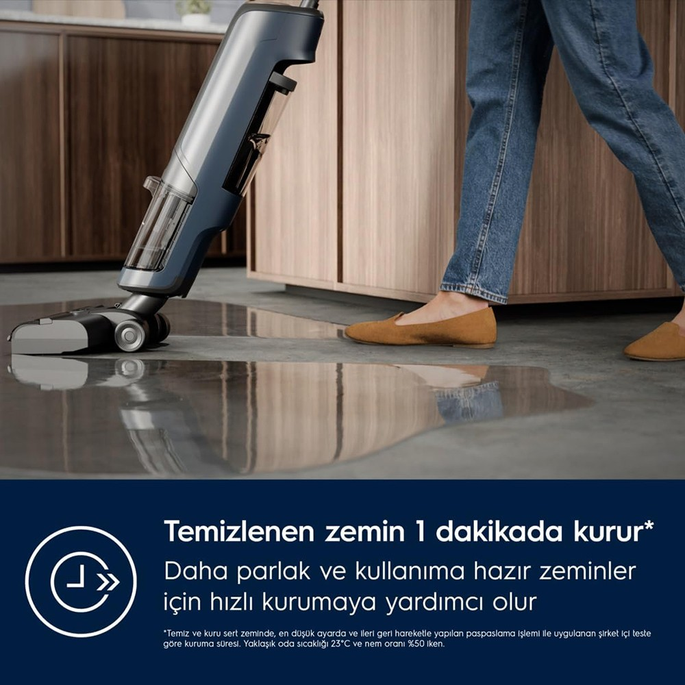 Un aspirateur balai bleu nettoie un sol mouillé, une personne en jean à proximité.