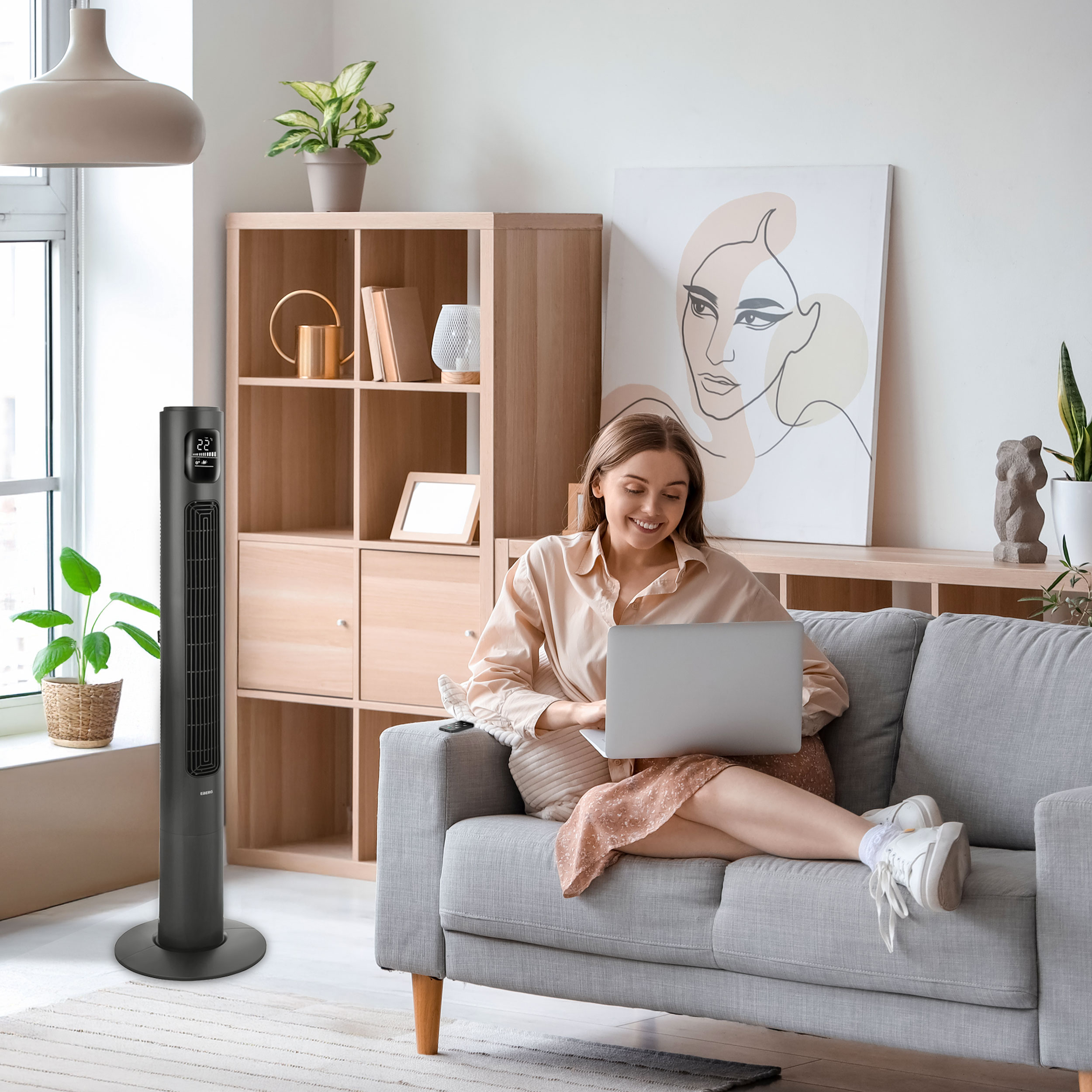 Kobieta siedzi na sofie, korzystając z laptopa. Obok stoi wentylator wieżowy.