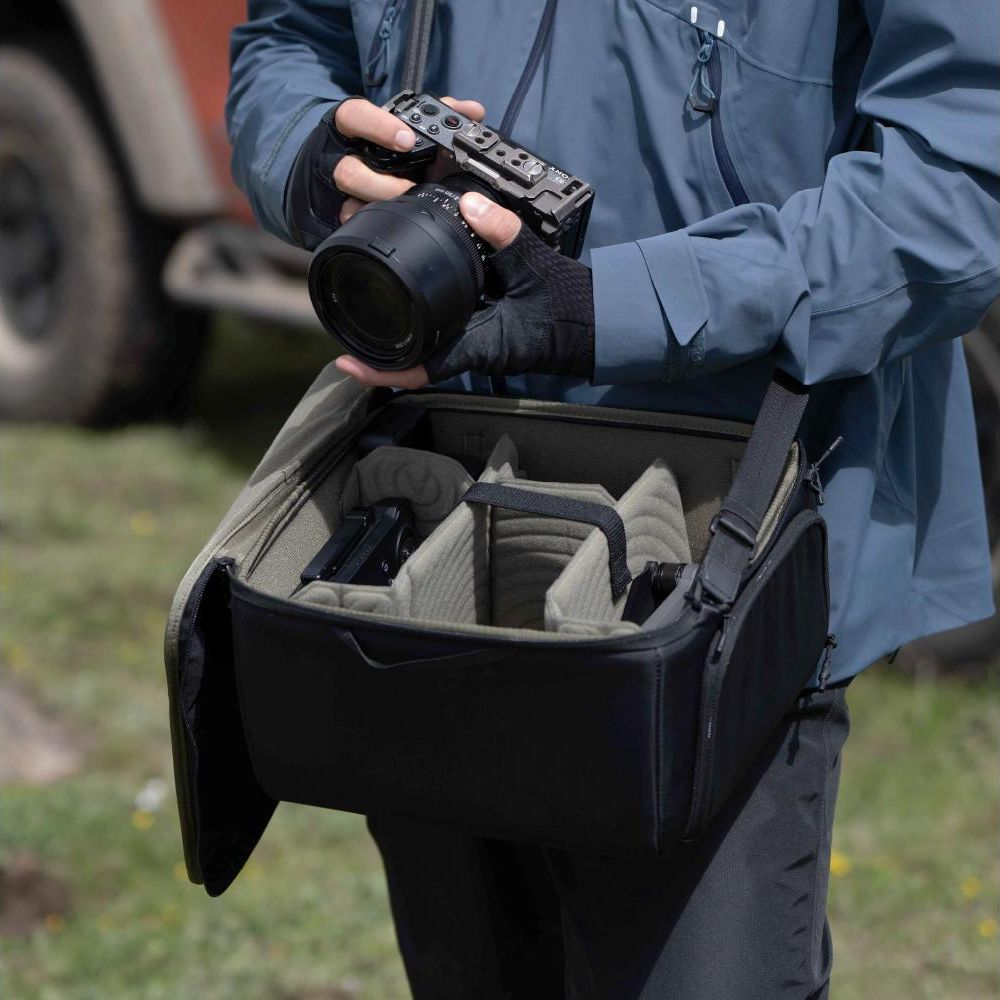 Persoon in blauwe jas houdt camera boven open tas met camera-uitrusting erin.