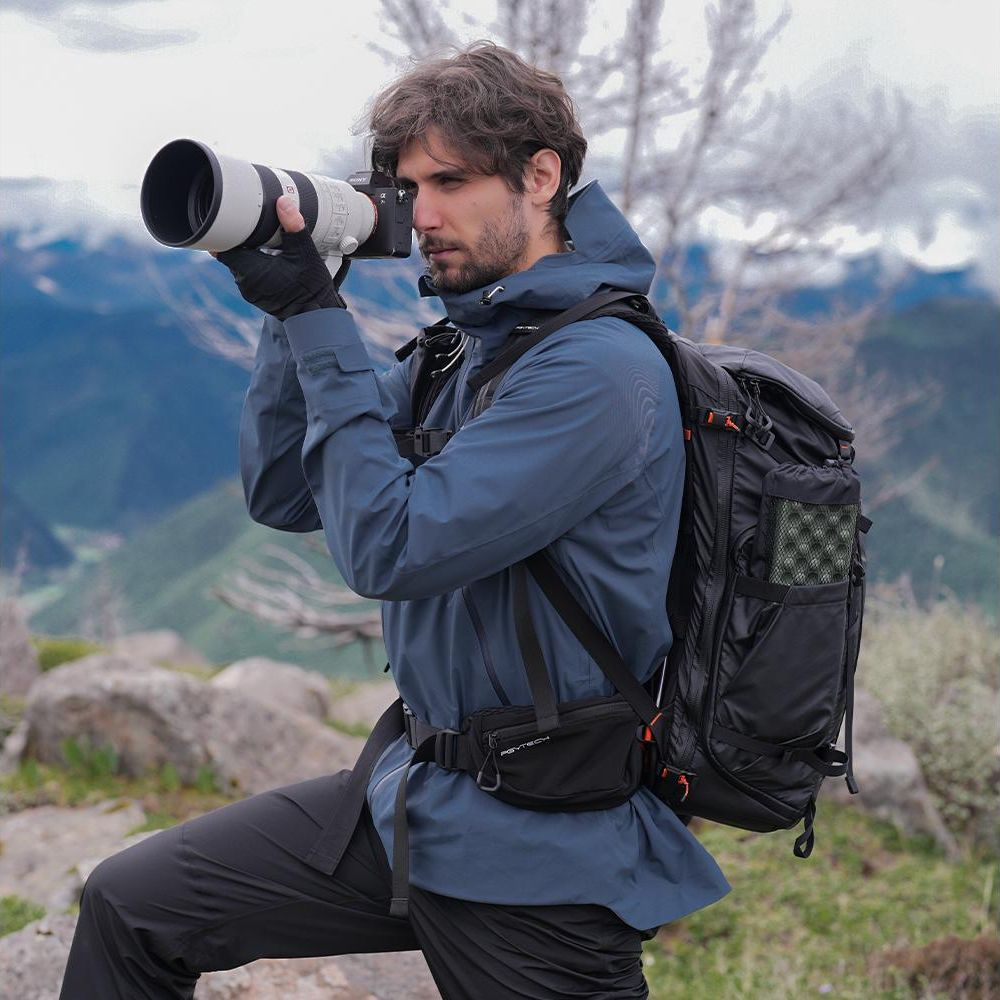 Een man in een blauwe jas met rugzak fotografeert in bergachtig landschap.