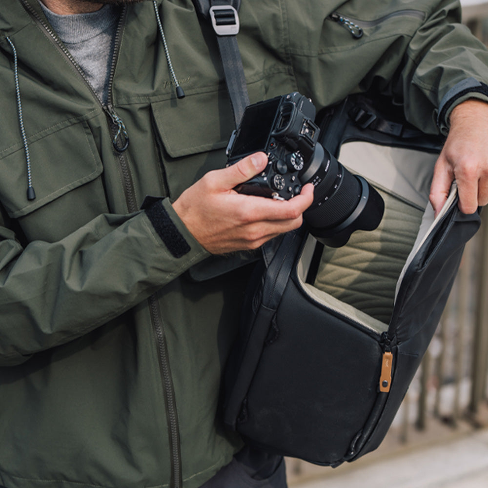 Homme en veste verte tient un appareil photo et ouvre un sac noir.