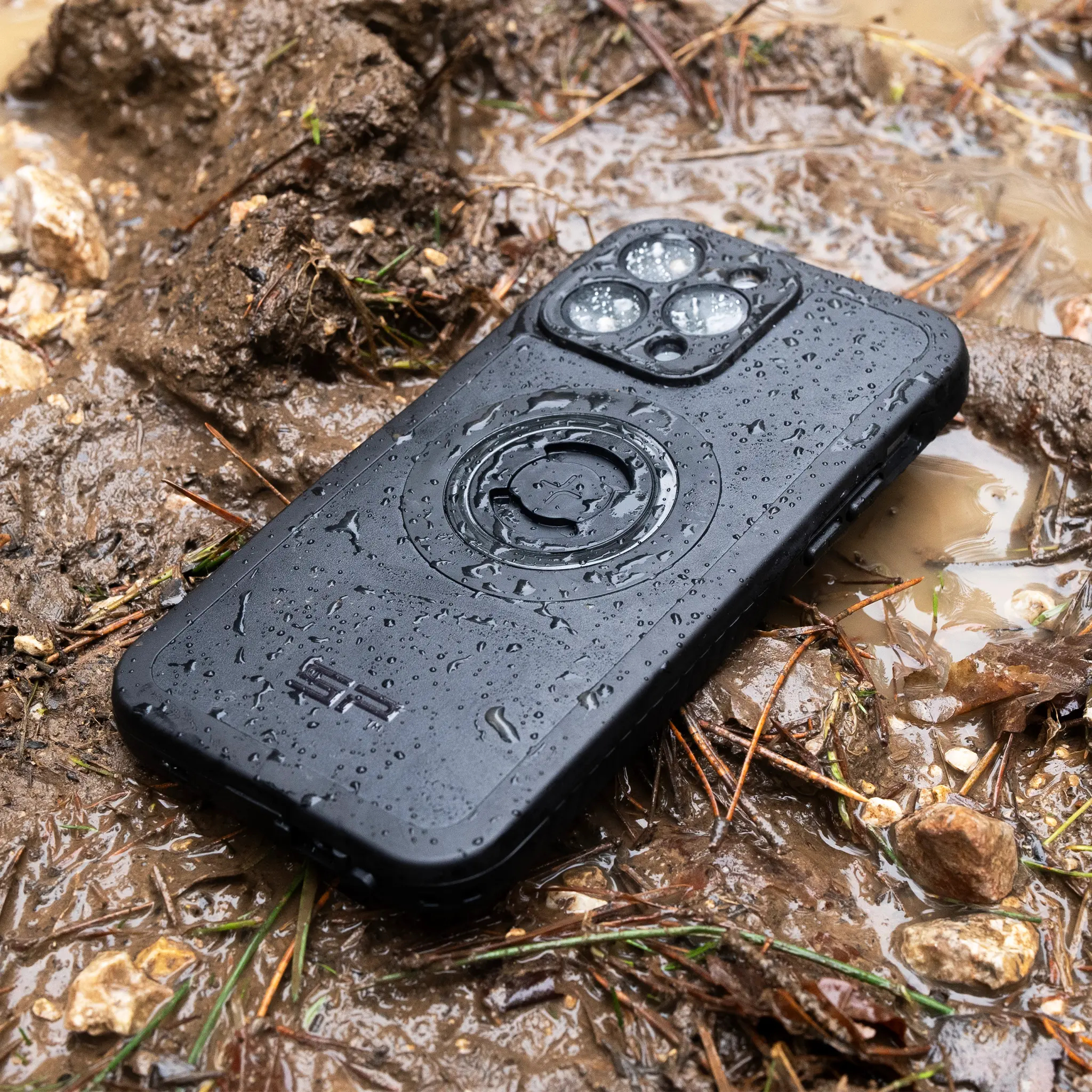 Ein schwarzes Smartphone mit drei Kameras liegt nass in Matsch und Ästen.