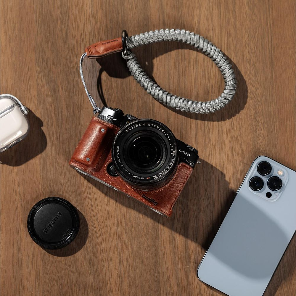 Custodia in pelle marrone, telefono, auricolari, tappo lente su tavolo legno.