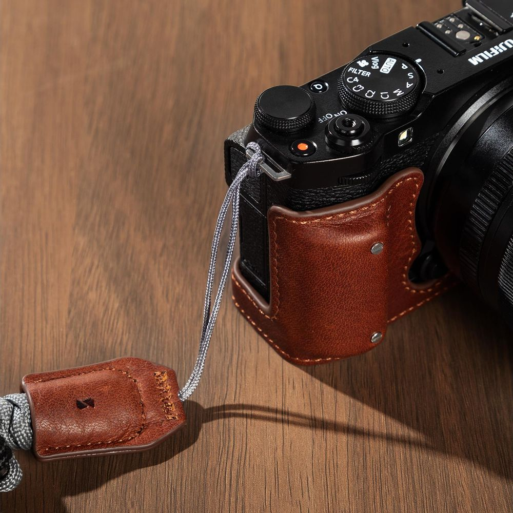 Fotocamera Fujifilm nera con mezza custodia in pelle marrone e cinturino da polso su legno.