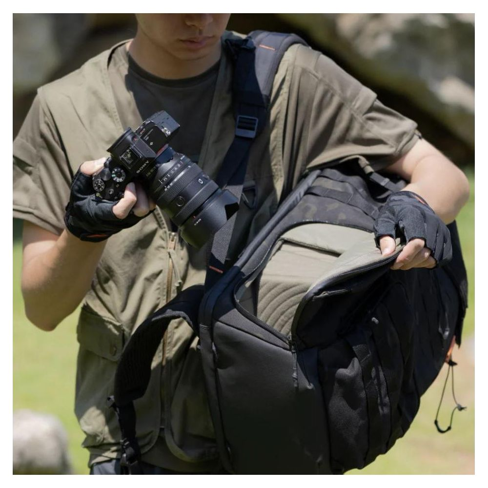 Persona in abbigliamento outdoor tiene fotocamera e apre zaino nero. Sfondo con erba verde.