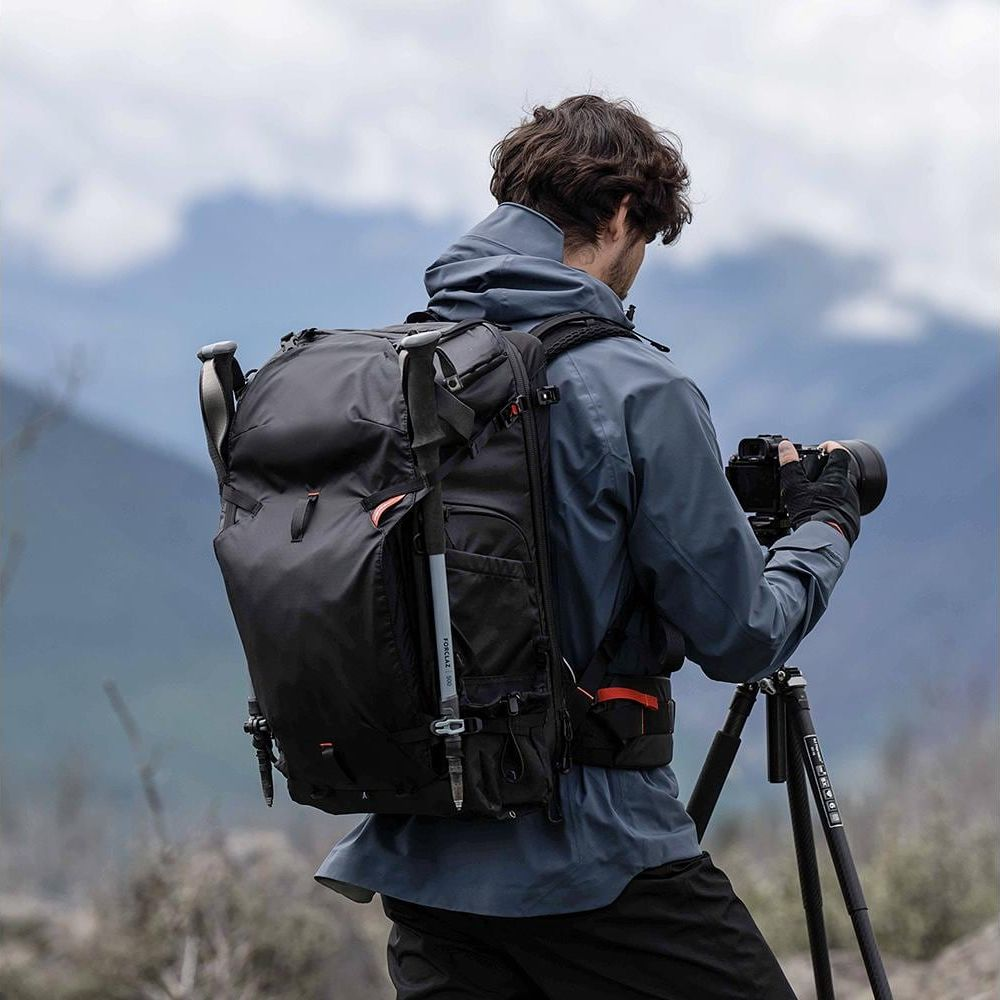 Homme avec sac à dos et appareil photo sur trépied dans paysage montagneux.