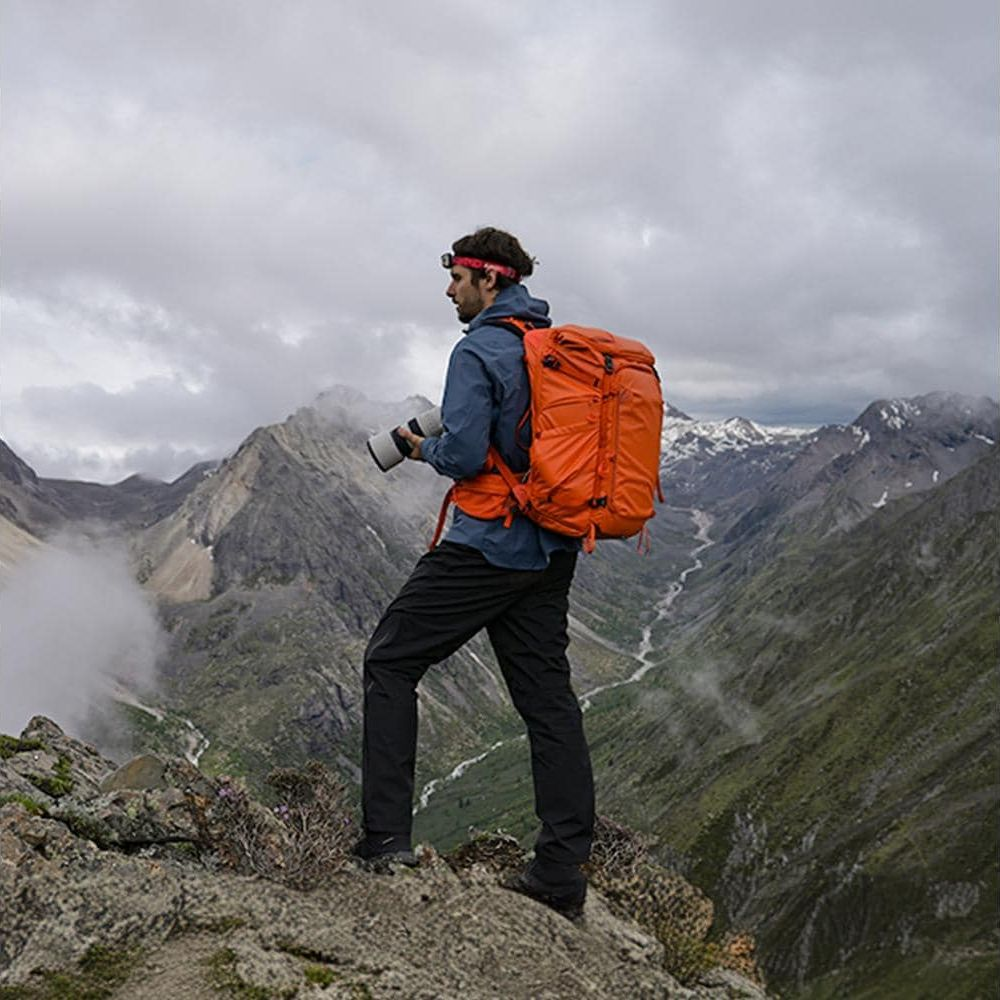 Ein Mann mit Rucksack und Kamera steht auf einem Felsen in den Bergen.