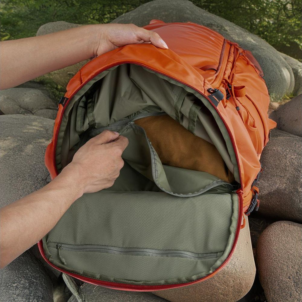 Eine Hand öffnet einen orangefarbenen Rucksack mit grünem Innenfutter, der auf Felsen liegt.