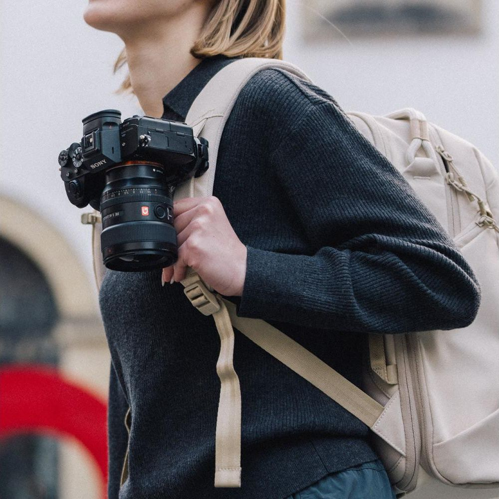 Een persoon draagt een zwarte Sony camera met lens en een beige rugzak.