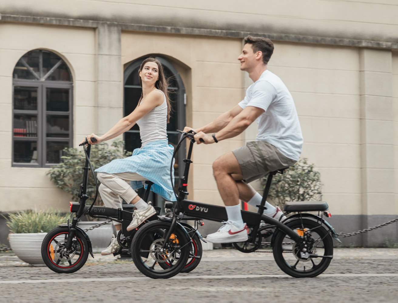 Un uomo e una donna vanno in bicicletta elettrica fianco a fianco davanti a un edificio.