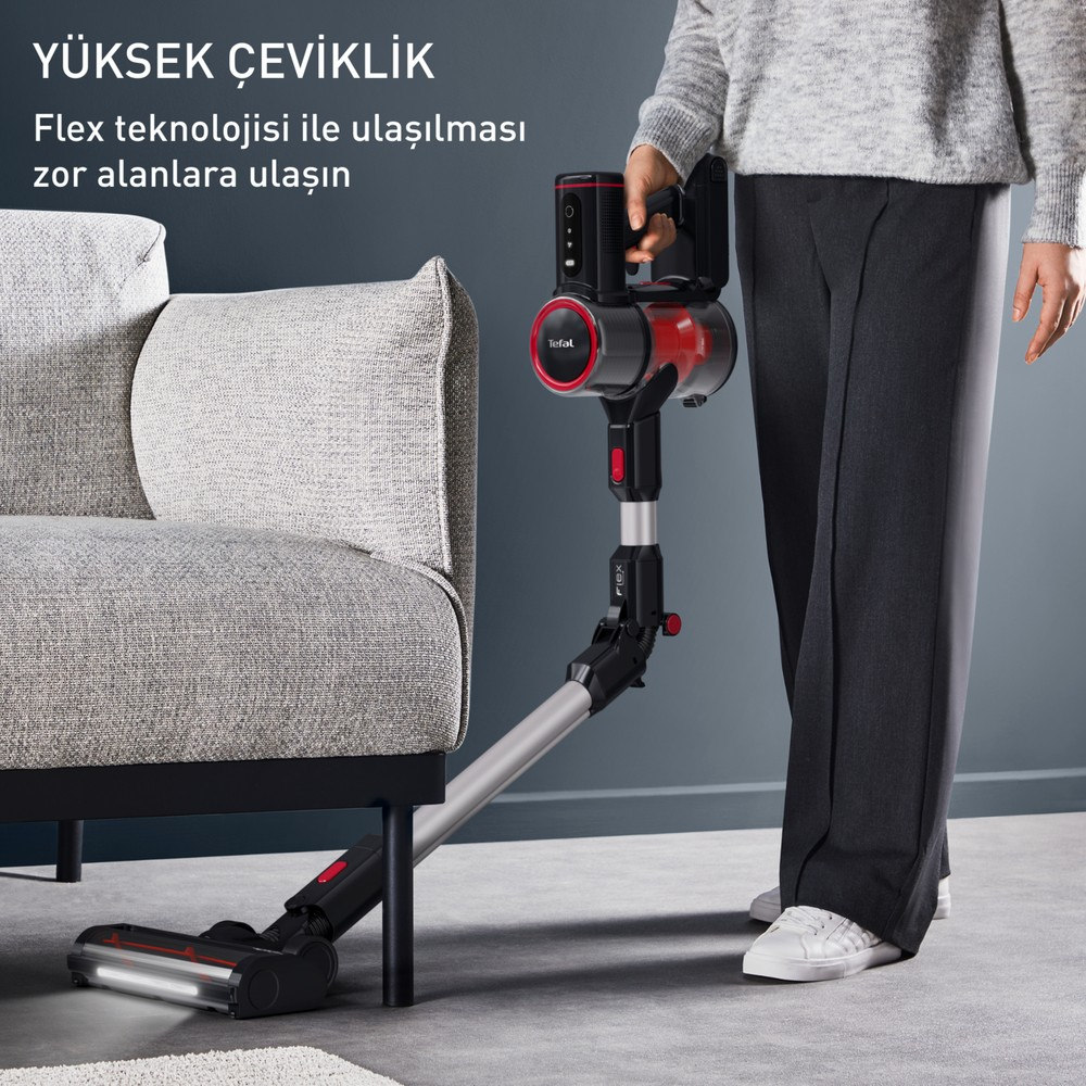 Kişi koltuğun altını temizlemek için kırmızı ve siyah dikey süpürge kullanıyor.