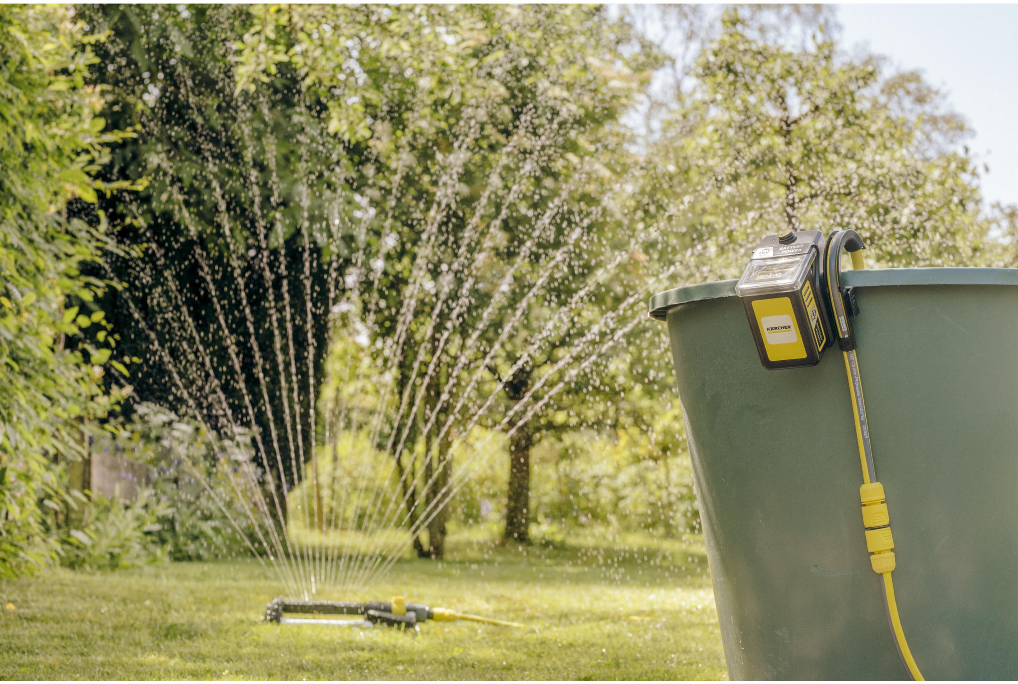 Un aspersor de césped riega el césped, un cubo verde con batería Karcher está al lado.