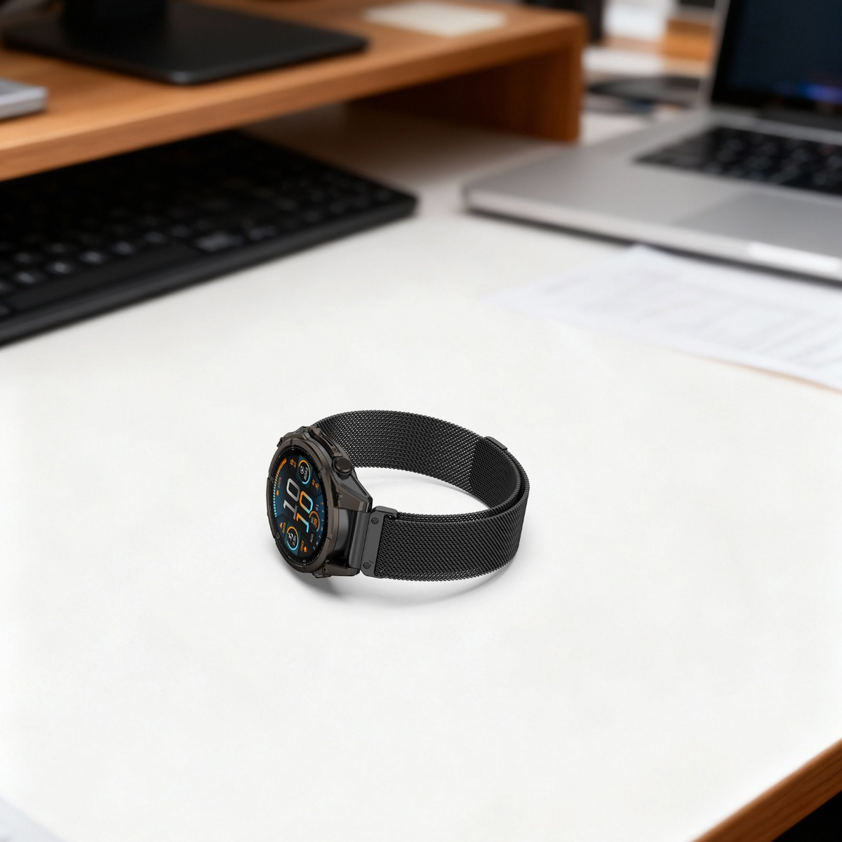 Uno smartwatch nero con cinturino in rete poggia su una scrivania bianca accanto a un laptop.