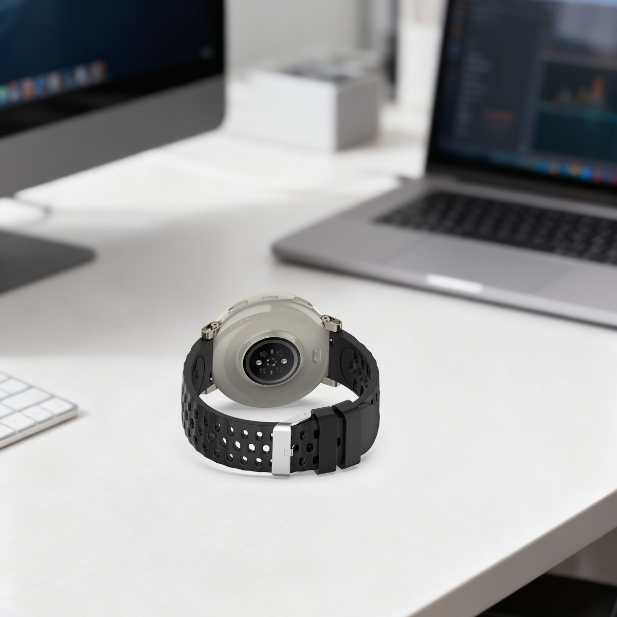 Eine Smartwatch mit schwarzem perforiertem Armband liegt auf einem weißen Schreibtisch neben einem Laptop und Monitor.