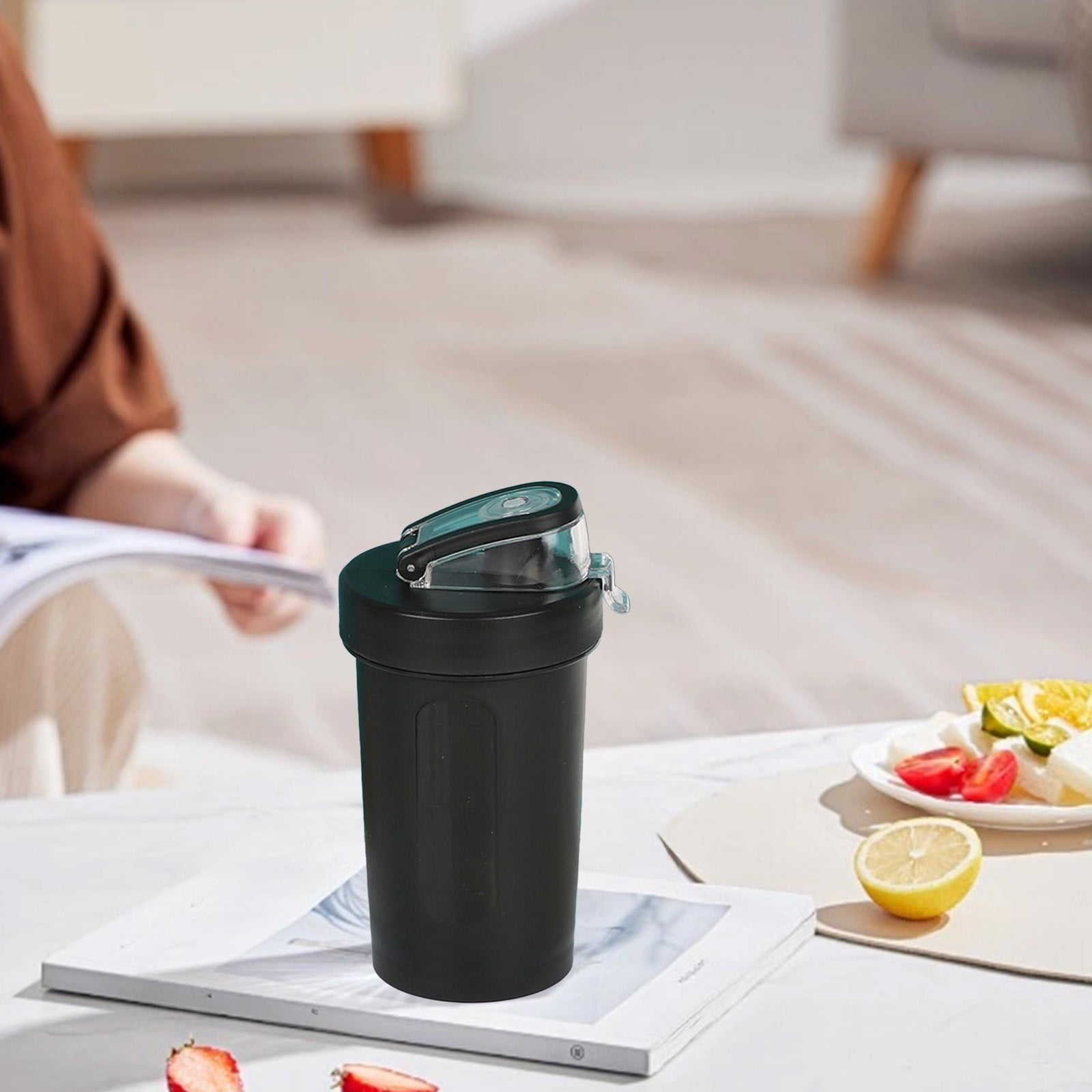 Schwarzer Shaker mit blauem Deckel auf einem Buch. Obst auf einem Teller im Hintergrund.