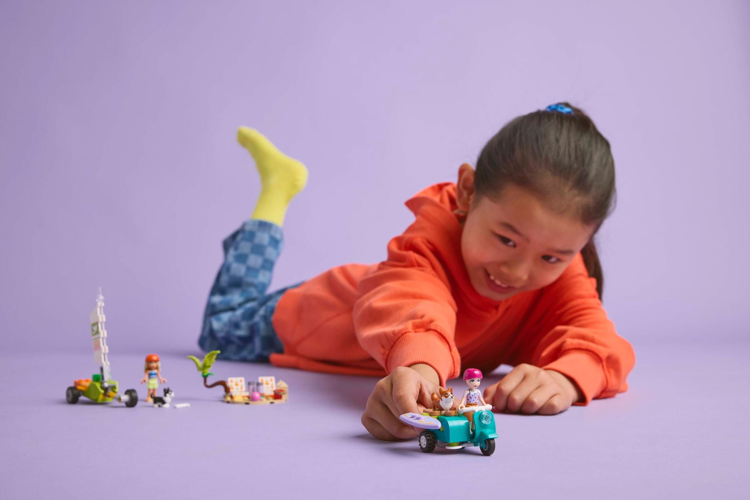 Kind spielt mit Legofiguren auf Roller und Segelwagen auf lila Hintergrund.