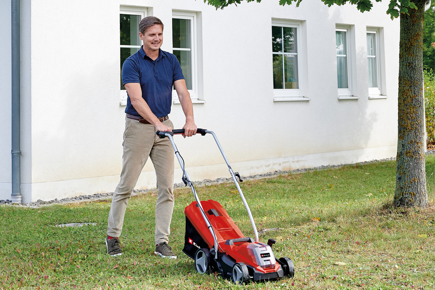 Ein Mann schiebt einen roten Rasenmäher im Garten vor einem weißen Haus.