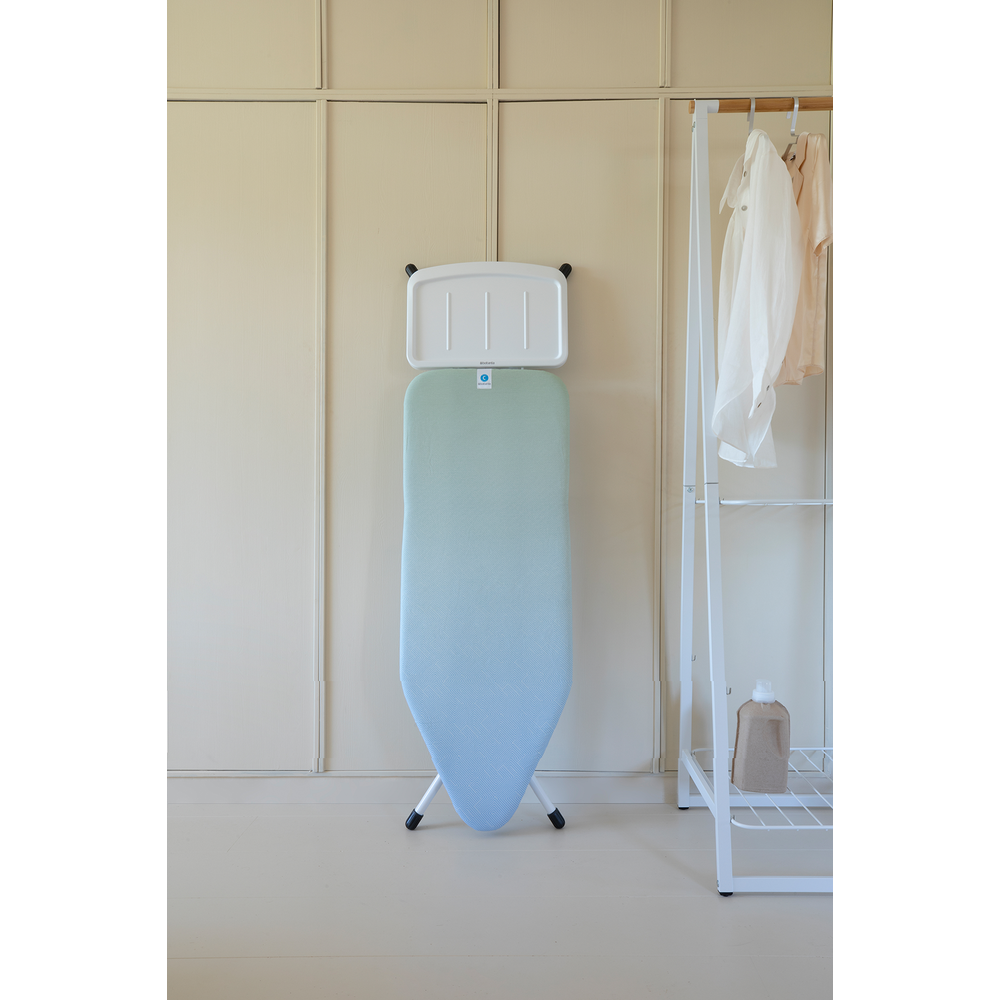 Een blauw-groen strijkplank staat tegen een muur. Kleding hangt aan een rek rechts.