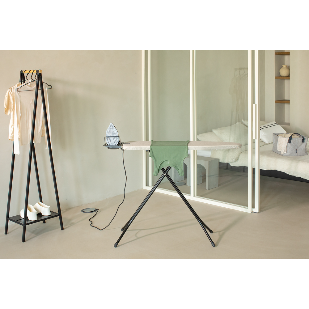 Un fer à repasser repose sur une planche à repasser avec une chemise verte. Porte-vêtements à proximité.