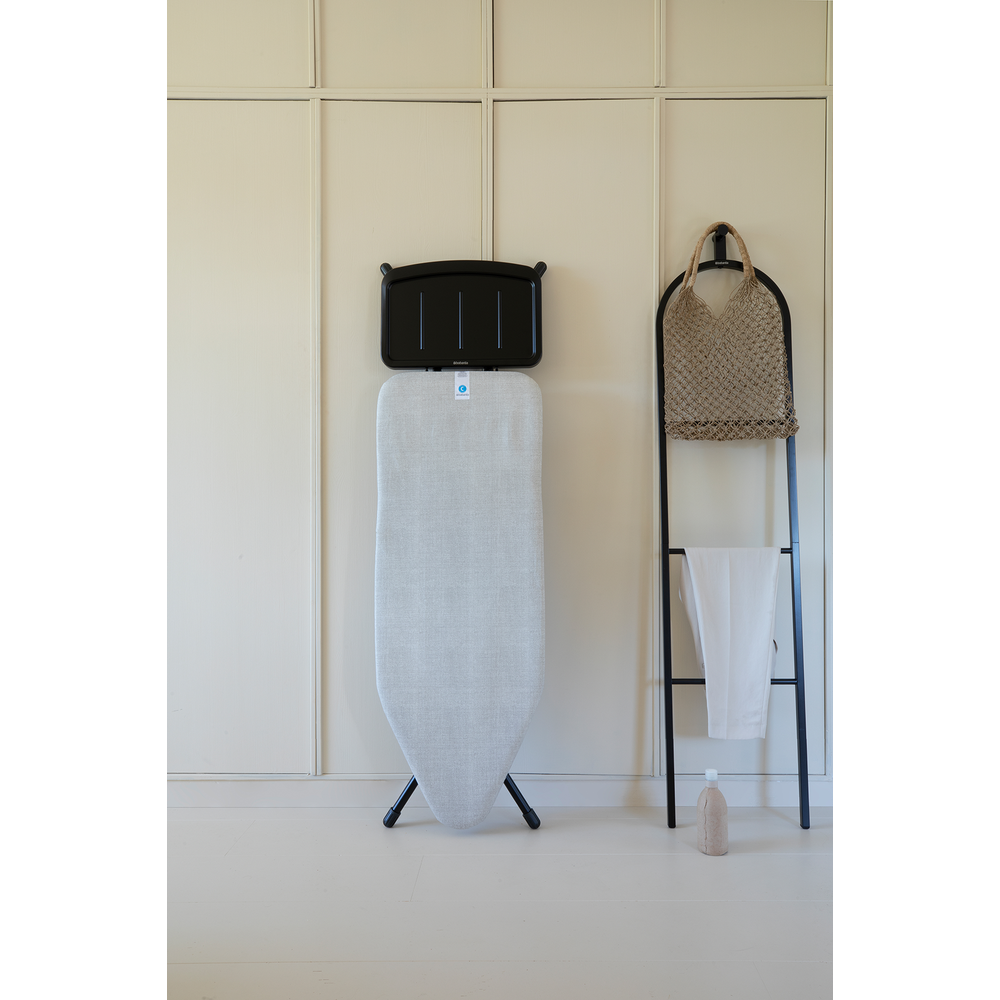 An ironing board with black holder, next to it a ladder rack with bag and cloth.