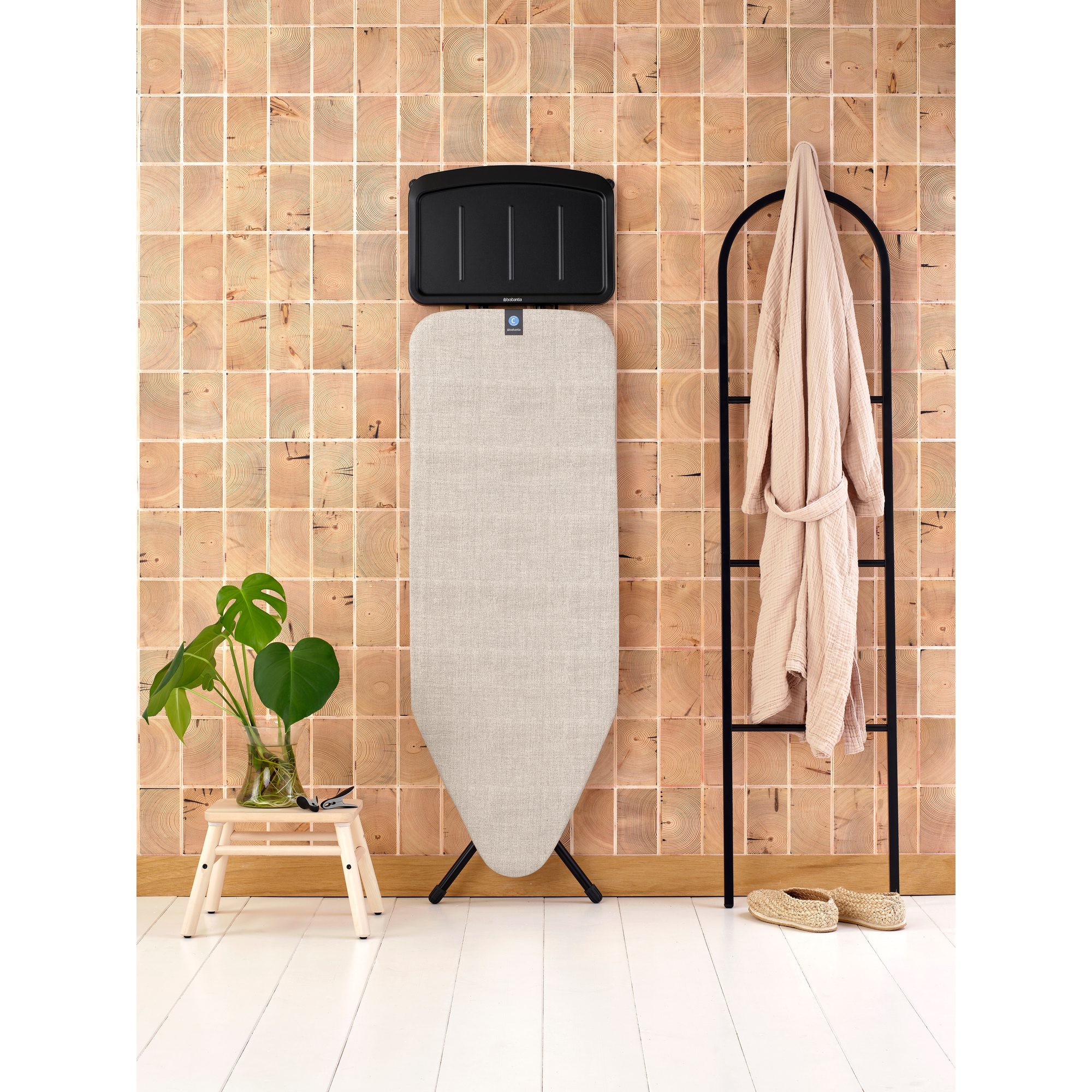 Ironing board with clothes rack, robe, plant, and stool.