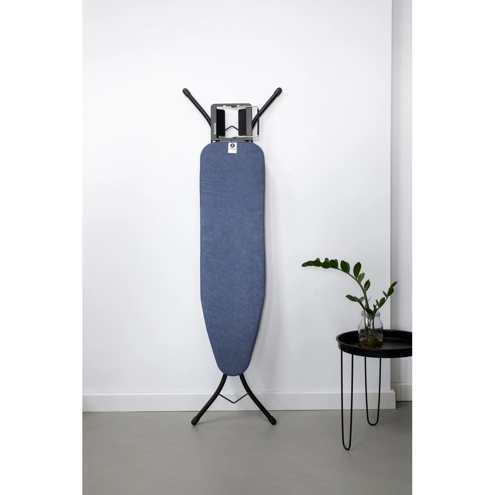 Une planche à repasser bleue se dresse contre un mur blanc à côté d'une petite table noire avec une plante.