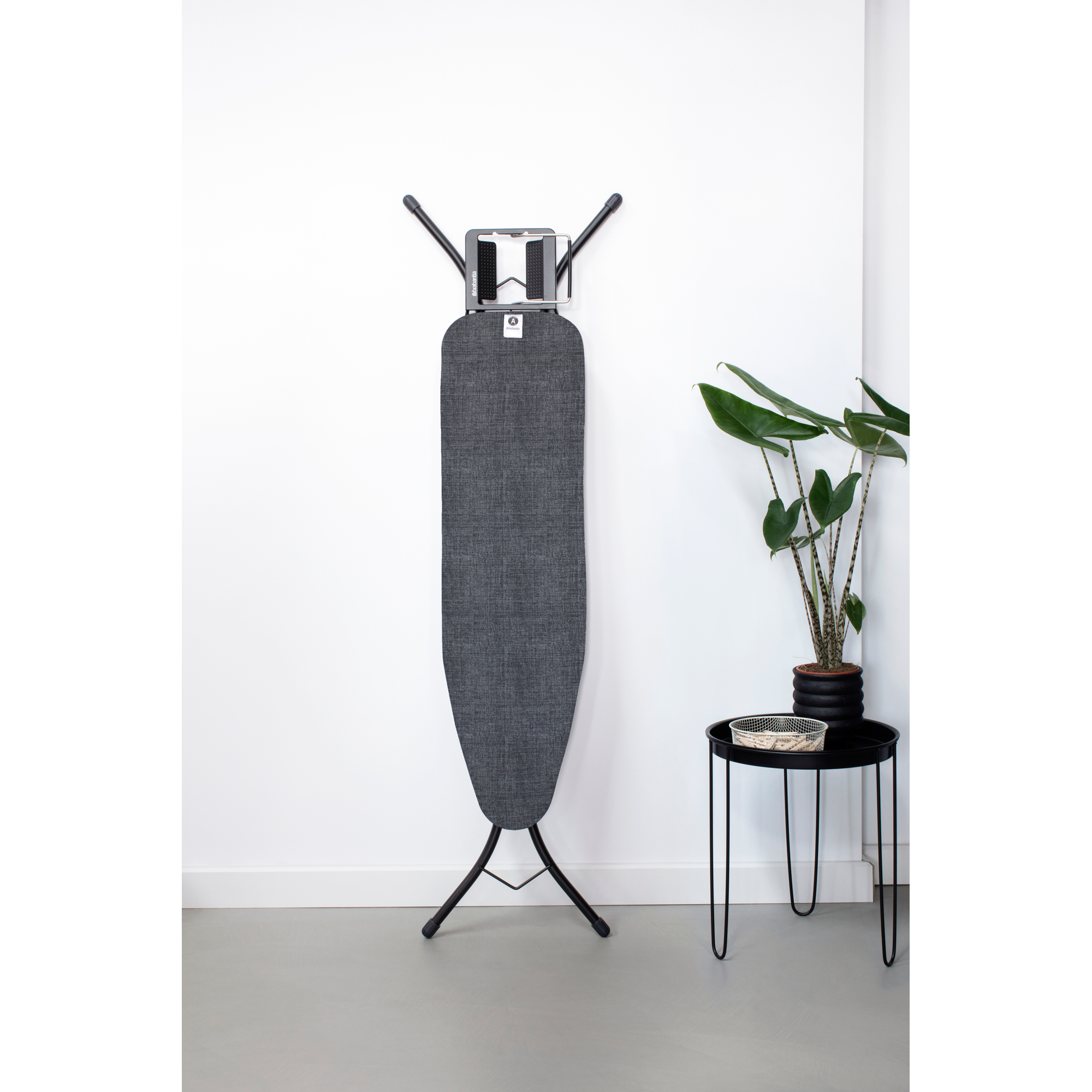 Une planche à repasser grise est debout contre un mur blanc à côté d'une plante.
