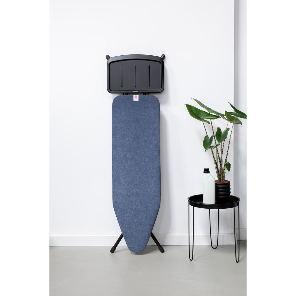 Une planche à repasser bleue est contre un mur blanc, à côté d'une petite table avec une plante.