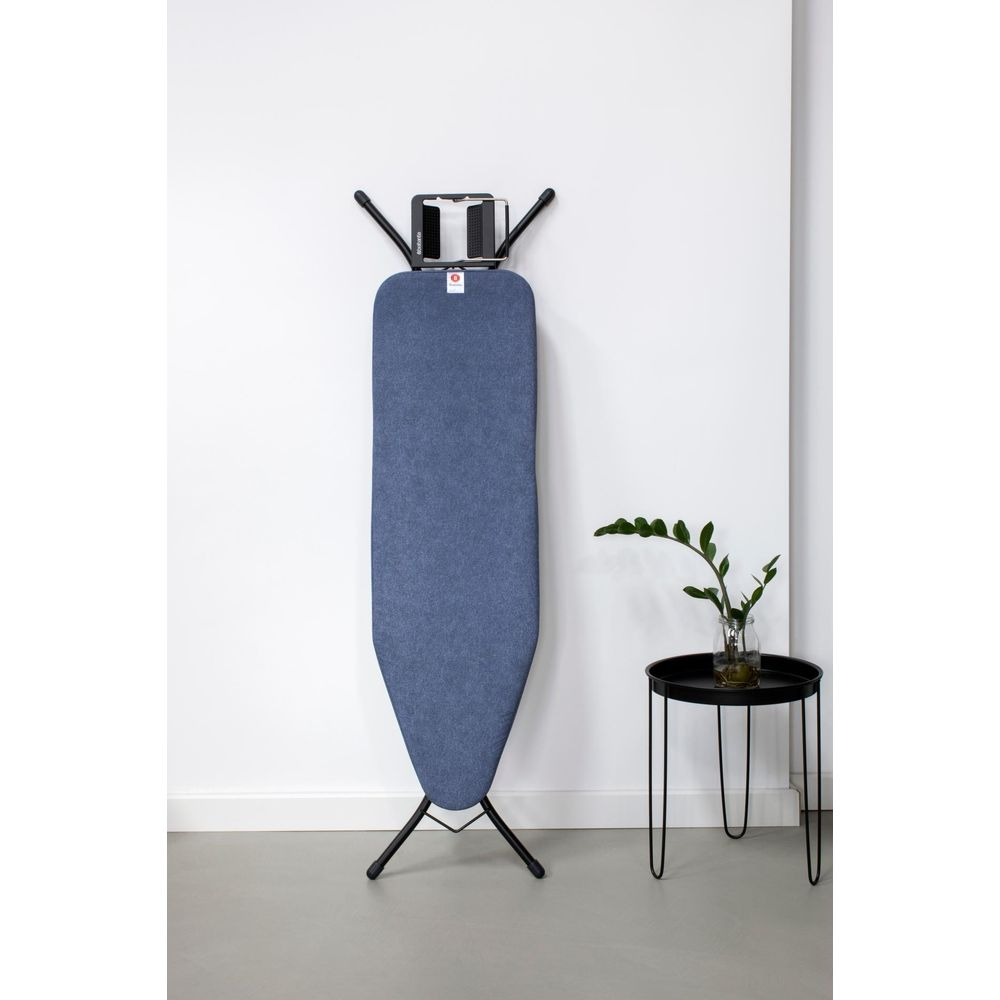 Une planche à repasser bleue est contre un mur blanc, avec une petite table noire et une plante.