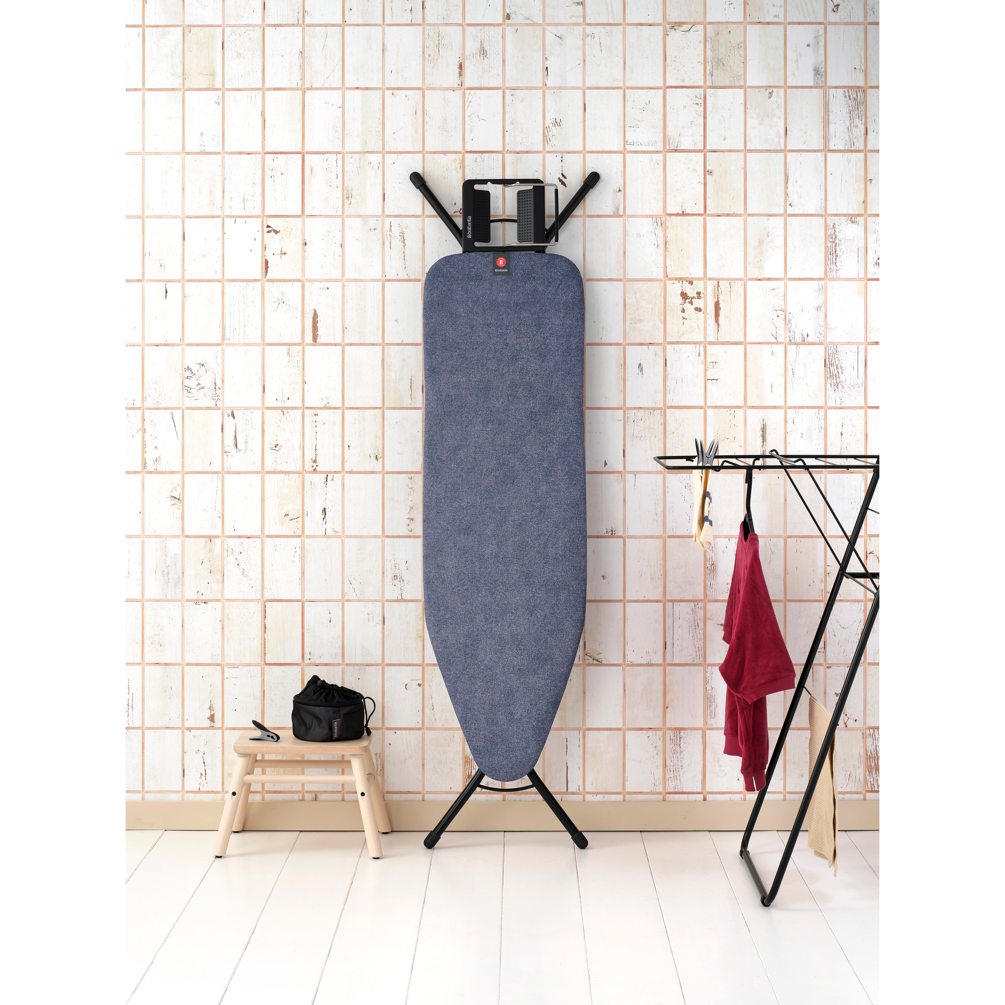 Une planche à repasser devant un mur carrelé. Tabouret avec sac à gauche, étendoir à linge à droite.