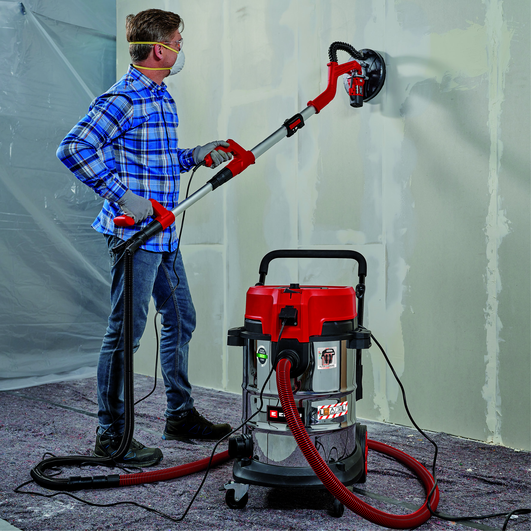 Mann schleift Wand mit langem Werkzeug und Staubsauger im Raum.