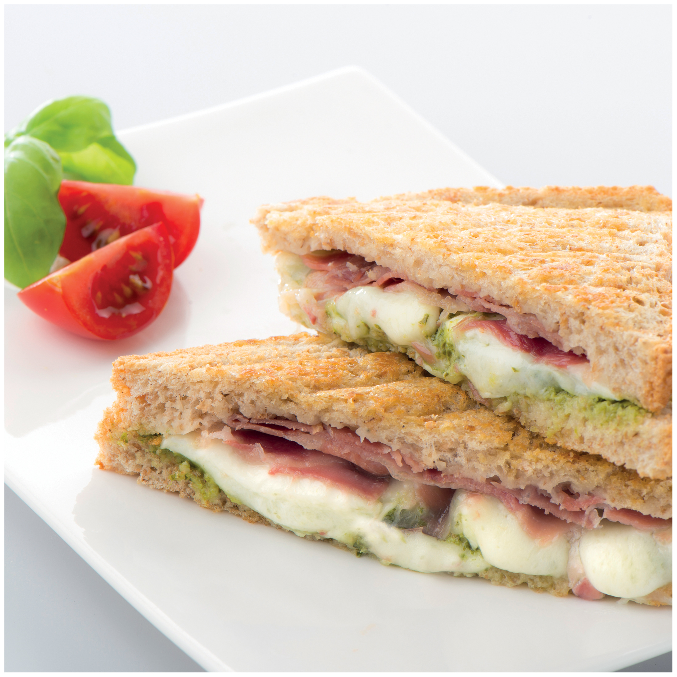 A sandwich with ham, mozzarella, and pesto on whole wheat toast, with tomatoes and basil.