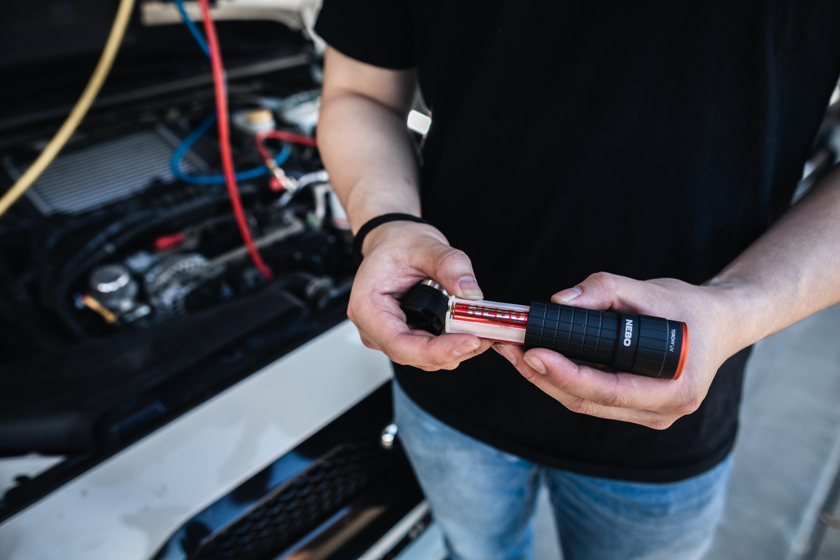 Persona inserisce batterie in una torcia UV nera, motore auto sullo sfondo.