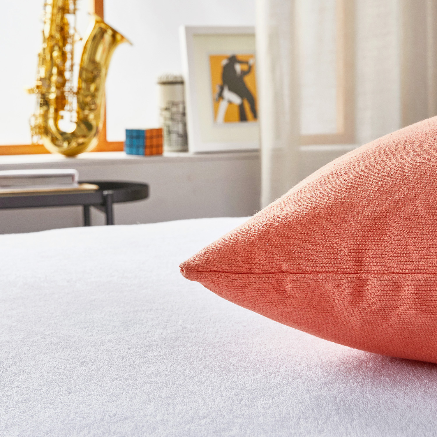 Un cojín coral en una cama blanca, con saxofón y cuadro al fondo.