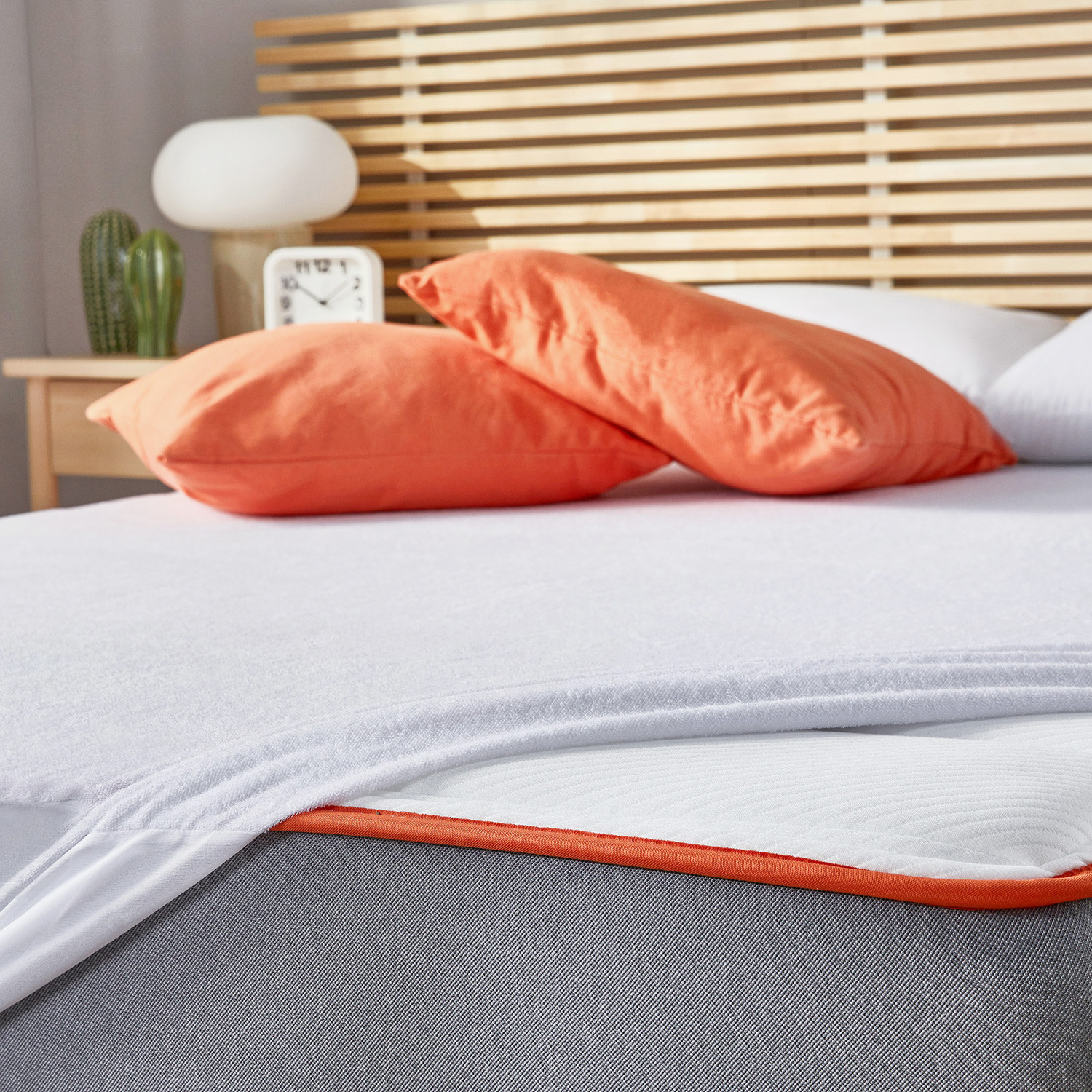 Una cama con almohadas naranjas y cabecero de madera. Lámpara y reloj visibles.