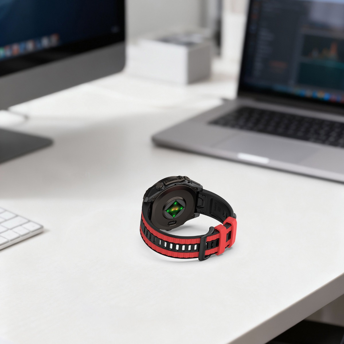 Schwarze Smartwatch mit rotem Armband auf einem Schreibtisch, Laptop und Monitor im Hintergrund.